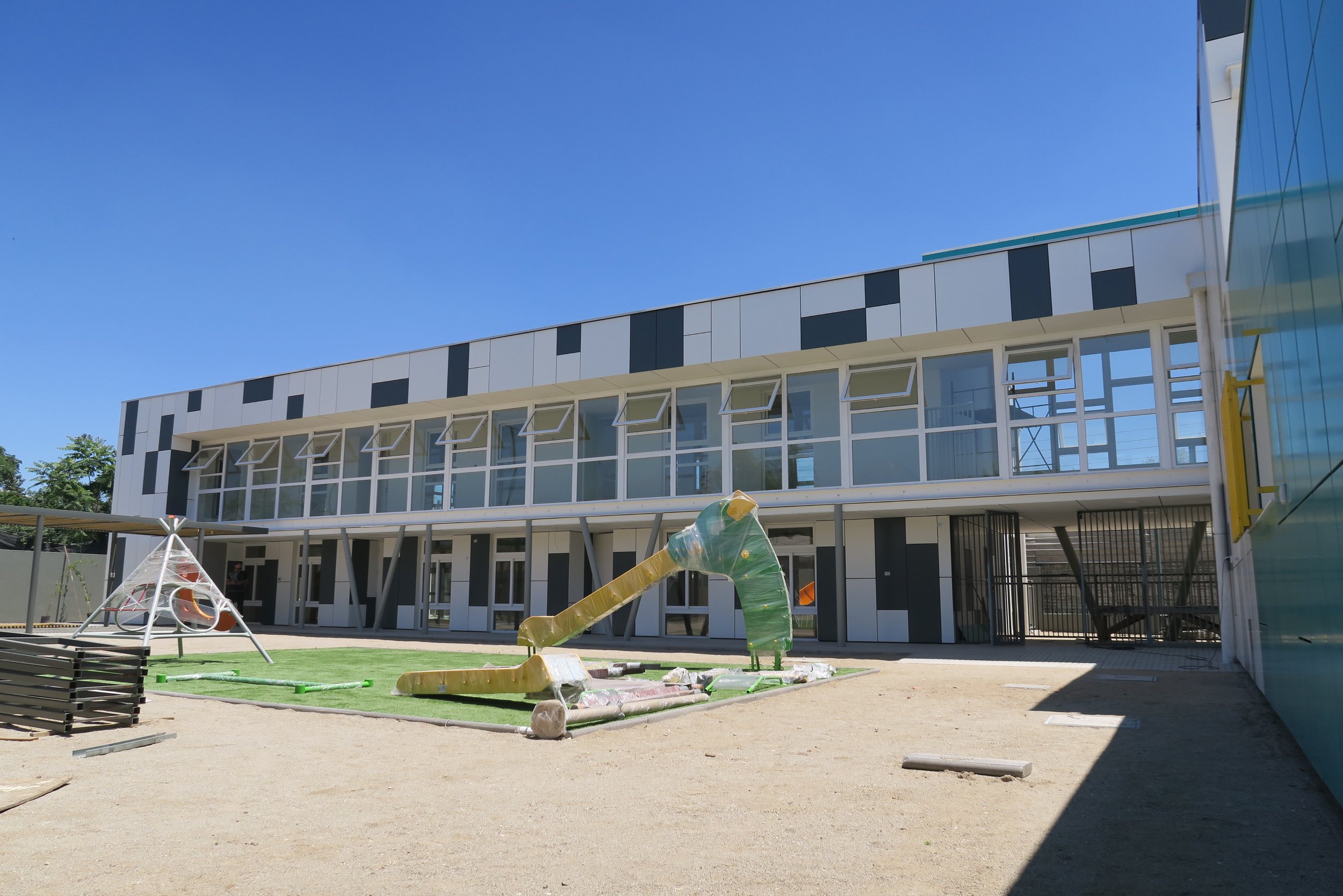 Edificio moderno en construcción con ventanas abiertas y área de juegos infantil en primer plano, cielo despejado.