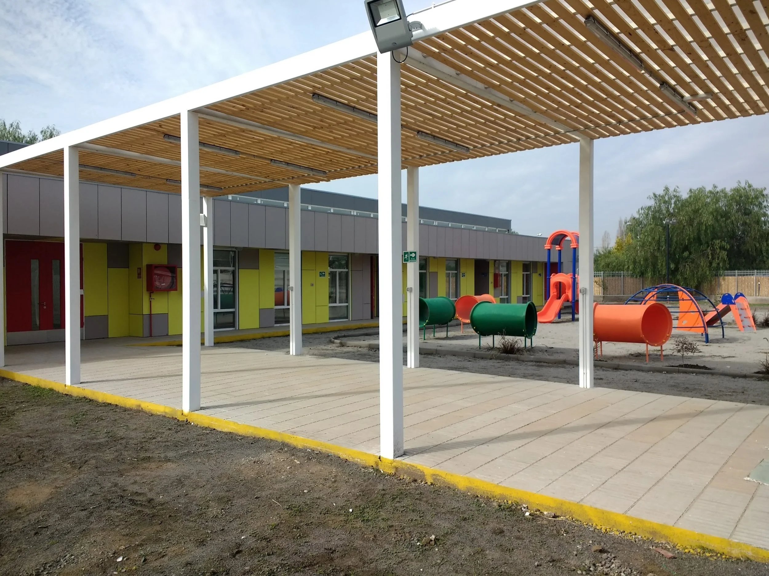 Espacio de recreo en una escuela con una estructura negra de madera, un suelo de baldosas y un área de arena con túneles y resbaladillas de plástico en colores naranja, verde y azul.