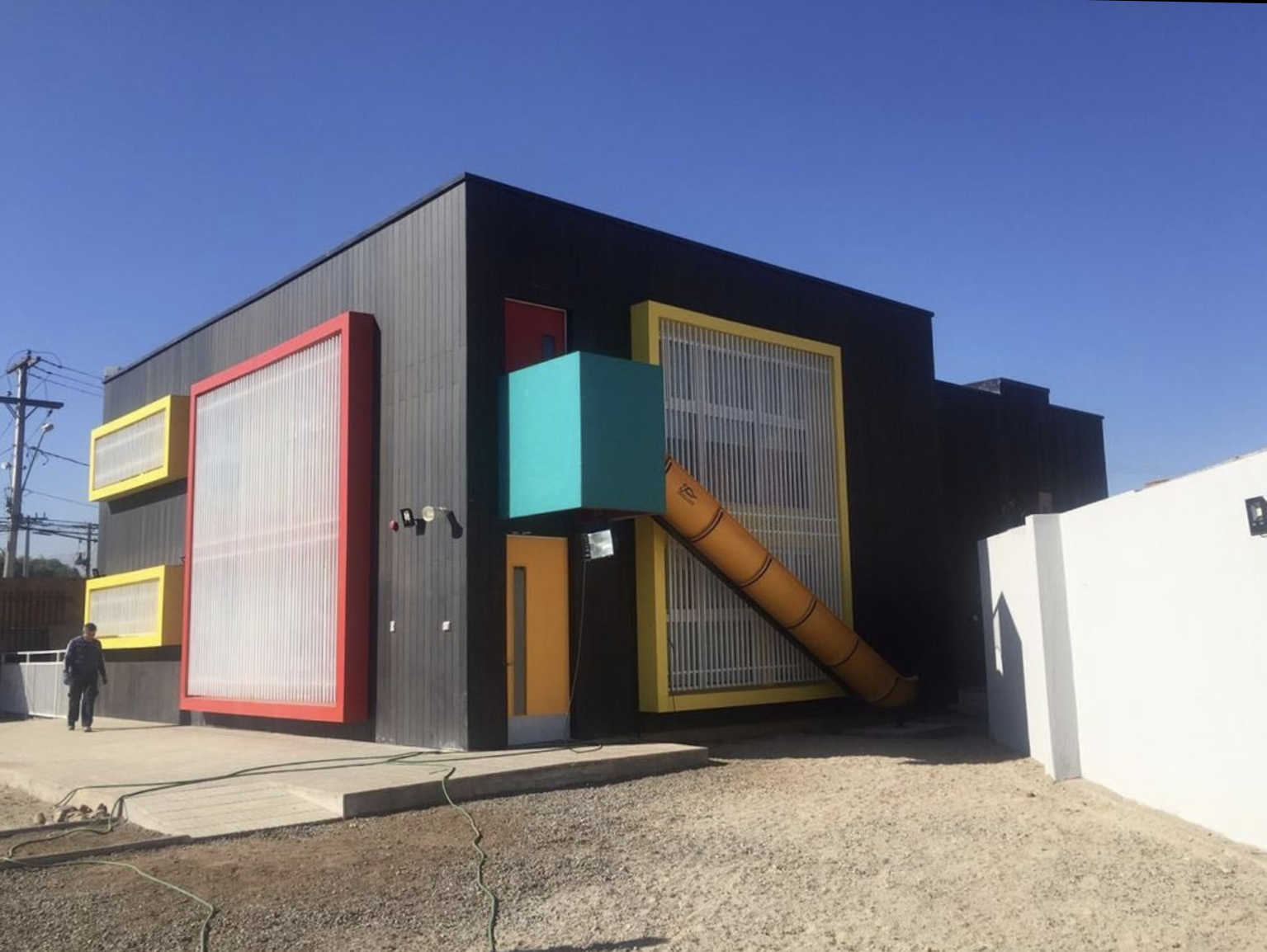 Edificio moderno con estructura colorida, ventanas de malla metálica y tubo de ventilación naranja, en un área exterior con camino de cemento y suelo de piedra.