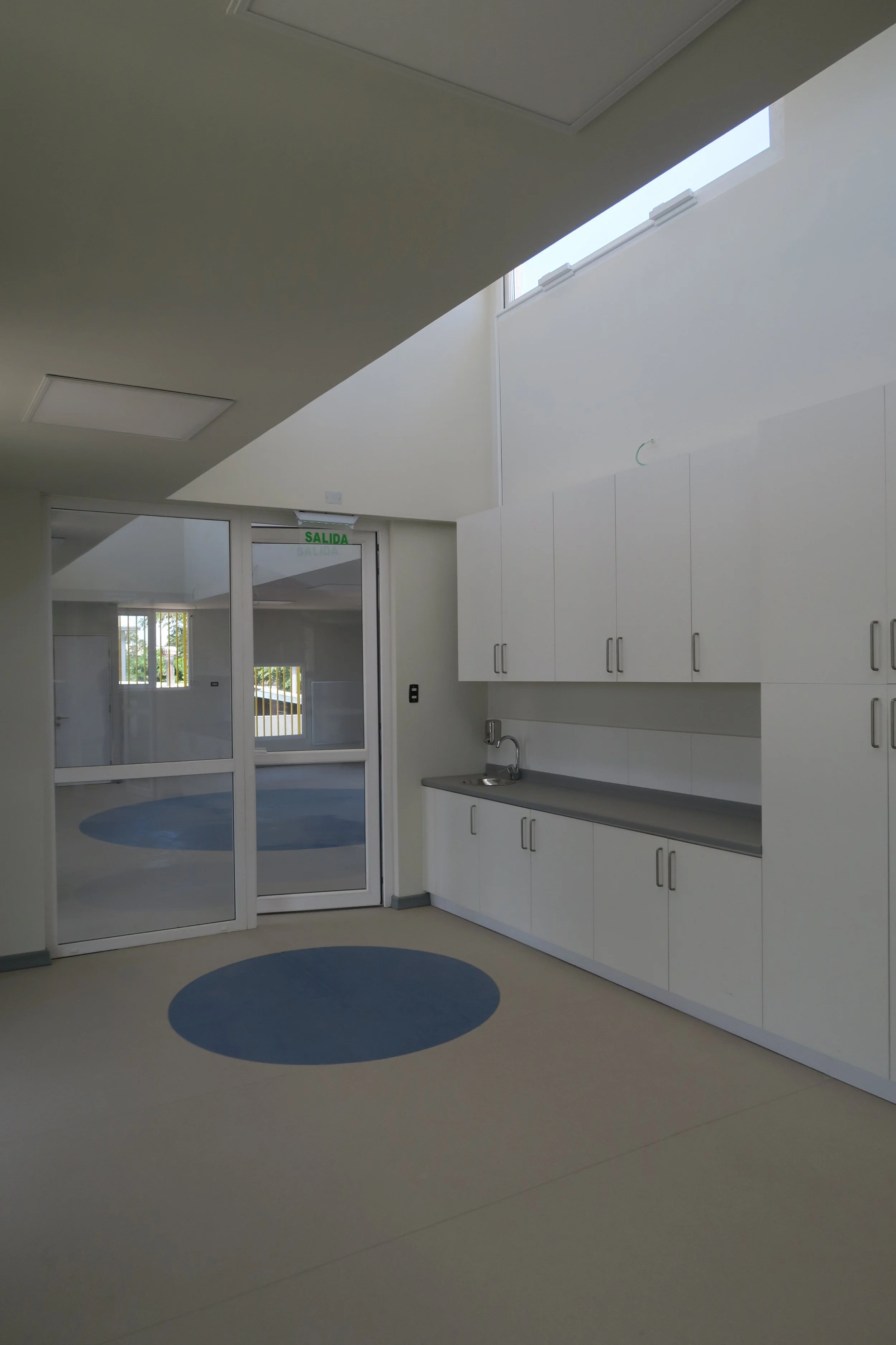 Interior de una sala blanca con gabinetes y un fregadero, puertas de vidrio hacia el exterior y ventanas altas que dejan entrar luz natural.