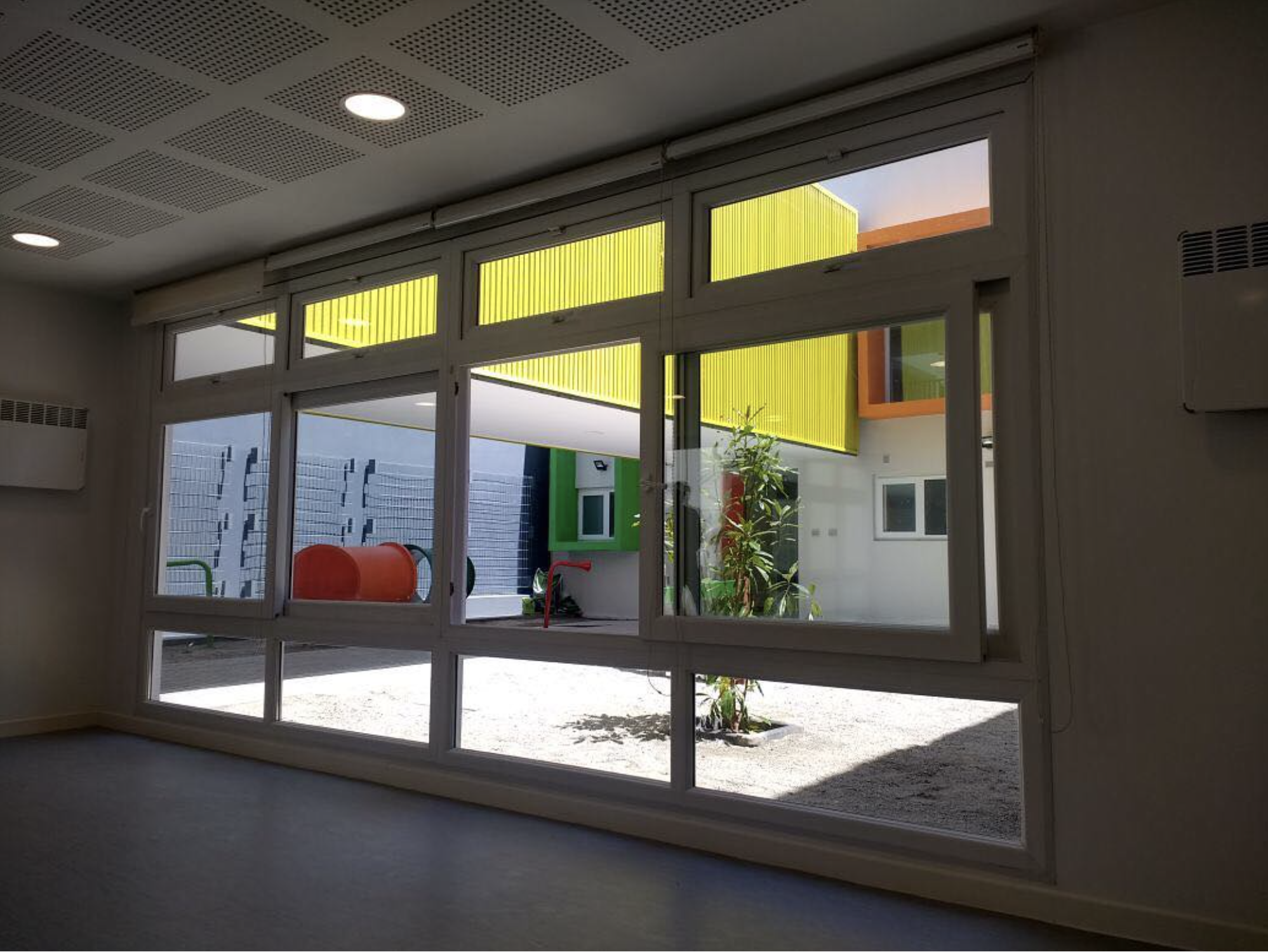 Vista desde dentro de una habitación con ventanas grandes que muestran un patio exterior con plantas, paredes coloridas y equipo de juegos para niños.