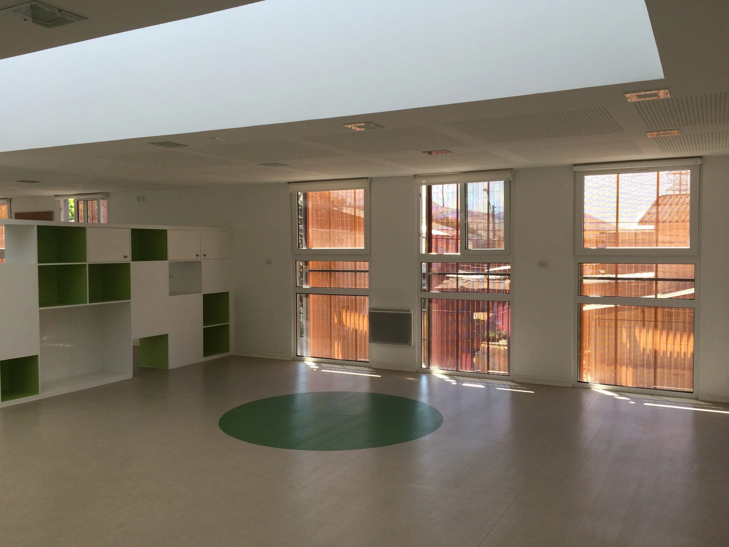Interior de una habitación vacía con ventanas grandes que dejan entrar luz natural y cortinas de color con patrón, con suelo de color claro y un espacio abierto, posiblemente un salón o sala de clases.