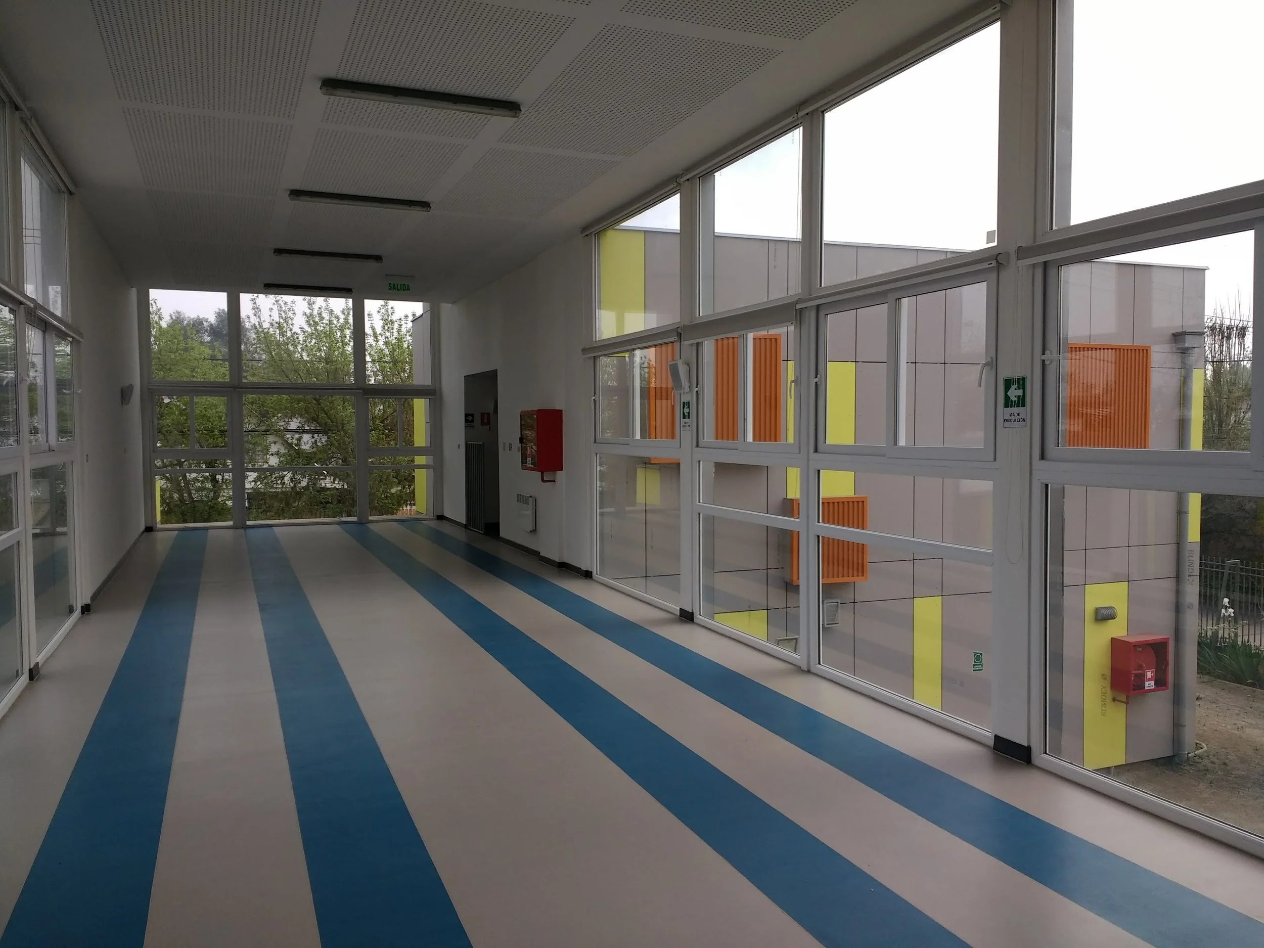 Interior de un pasillo de un edificio con paredes de vidrio y puertas de metal, piso con líneas azules y grises, ventanas grandes que muestran árboles y un cielo nublado, señal de salida y extintor de incendios visibles.
