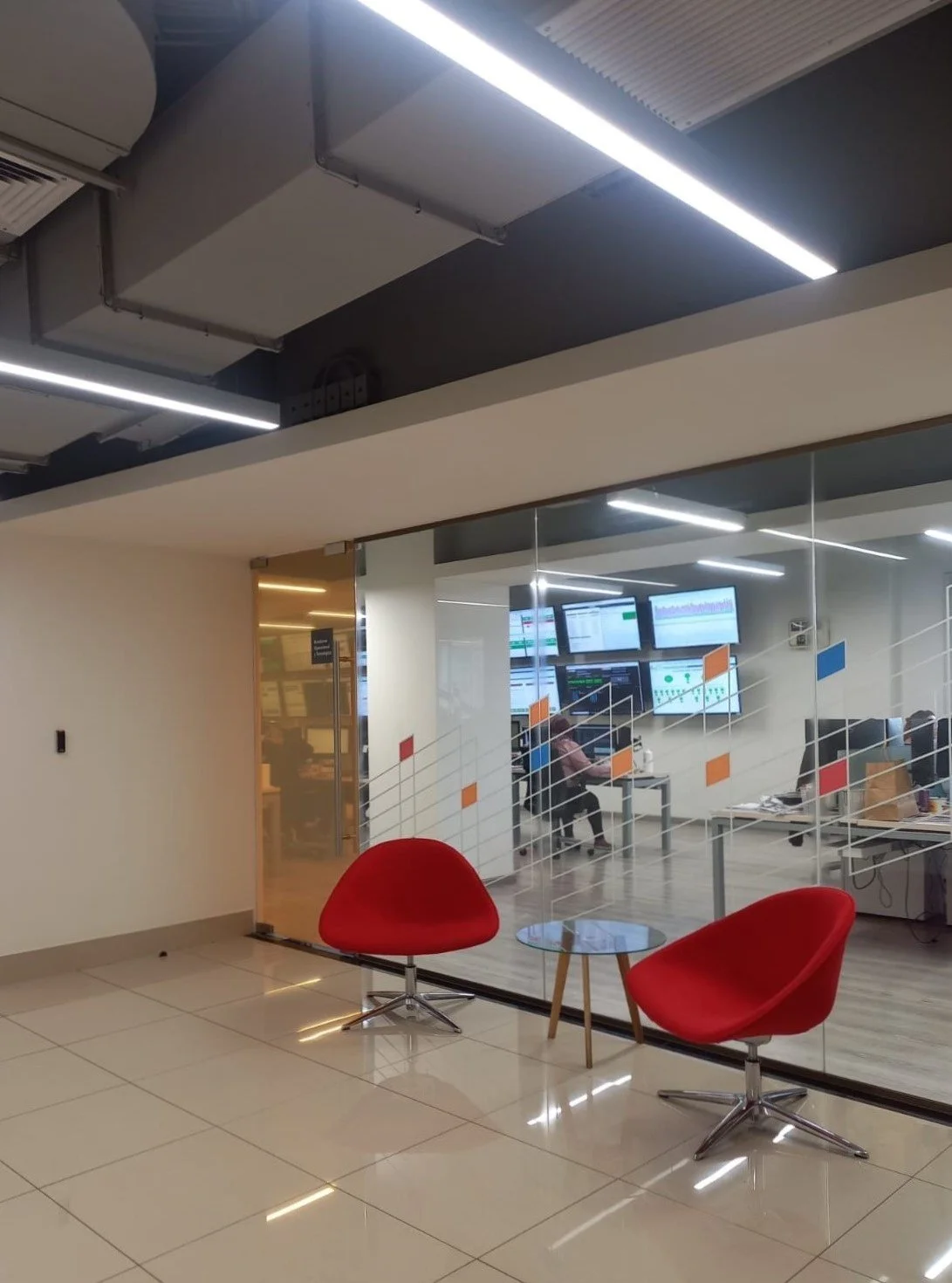 Vista de una sala de espera con dos sillones rojos y una mesa de centro, detrás un cristal que separa hacia una oficina con estaciones de trabajo y varias pantallas, en un ambiente moderno con iluminación
