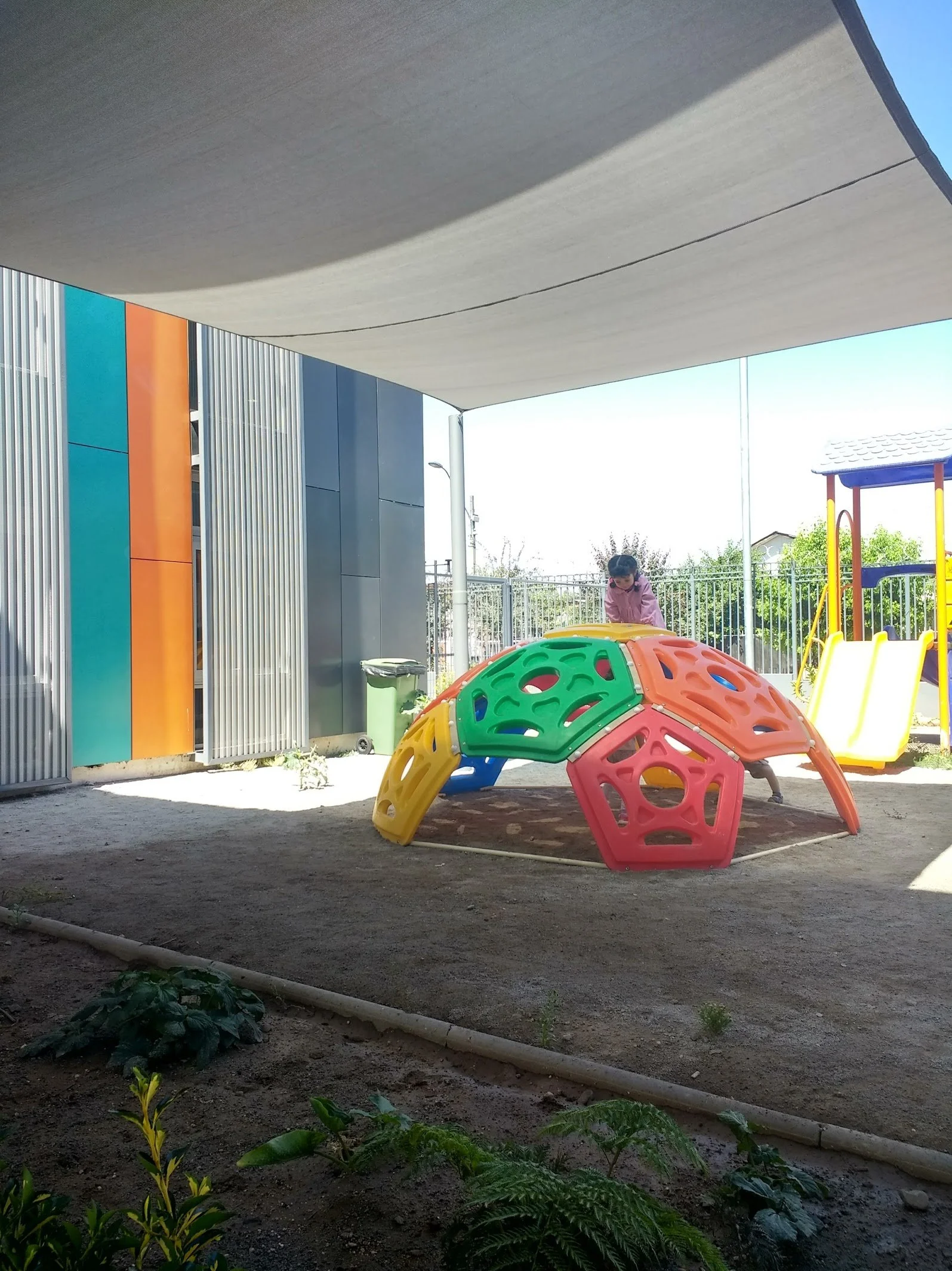 Niña jugando en un área infantil al aire libre con estructuras de plástico coloridas, bajo sombra de un techo y con un edificio moderno a un lado.