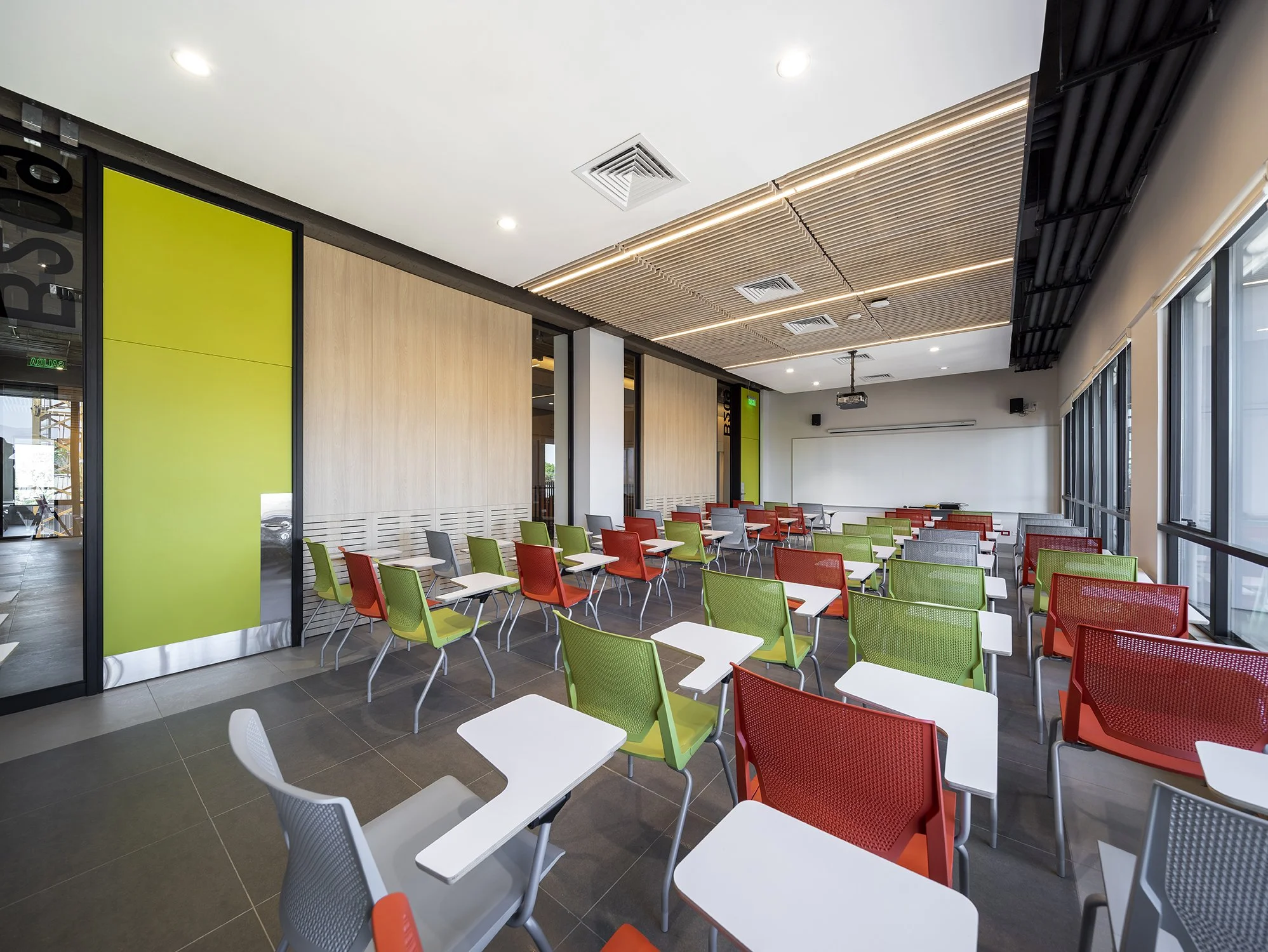 Aula moderna con sillas verdes, rojas y gris en filas, escritorio con computadora y proyector en el fondo, ventanas grandes y techo con paneles de madera.