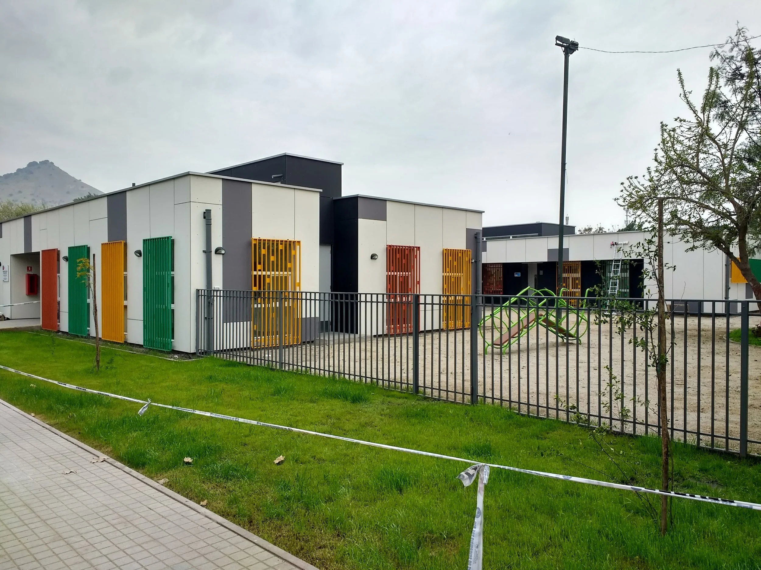 Edificio de estructura moderna con paredes blancas y detalles en colores rojo, verde y amarillo en las ventanas, cercado de hierro negro, árboles y área de juegos infantiles en primer plano, y montañas al fondo, en un día nublado.