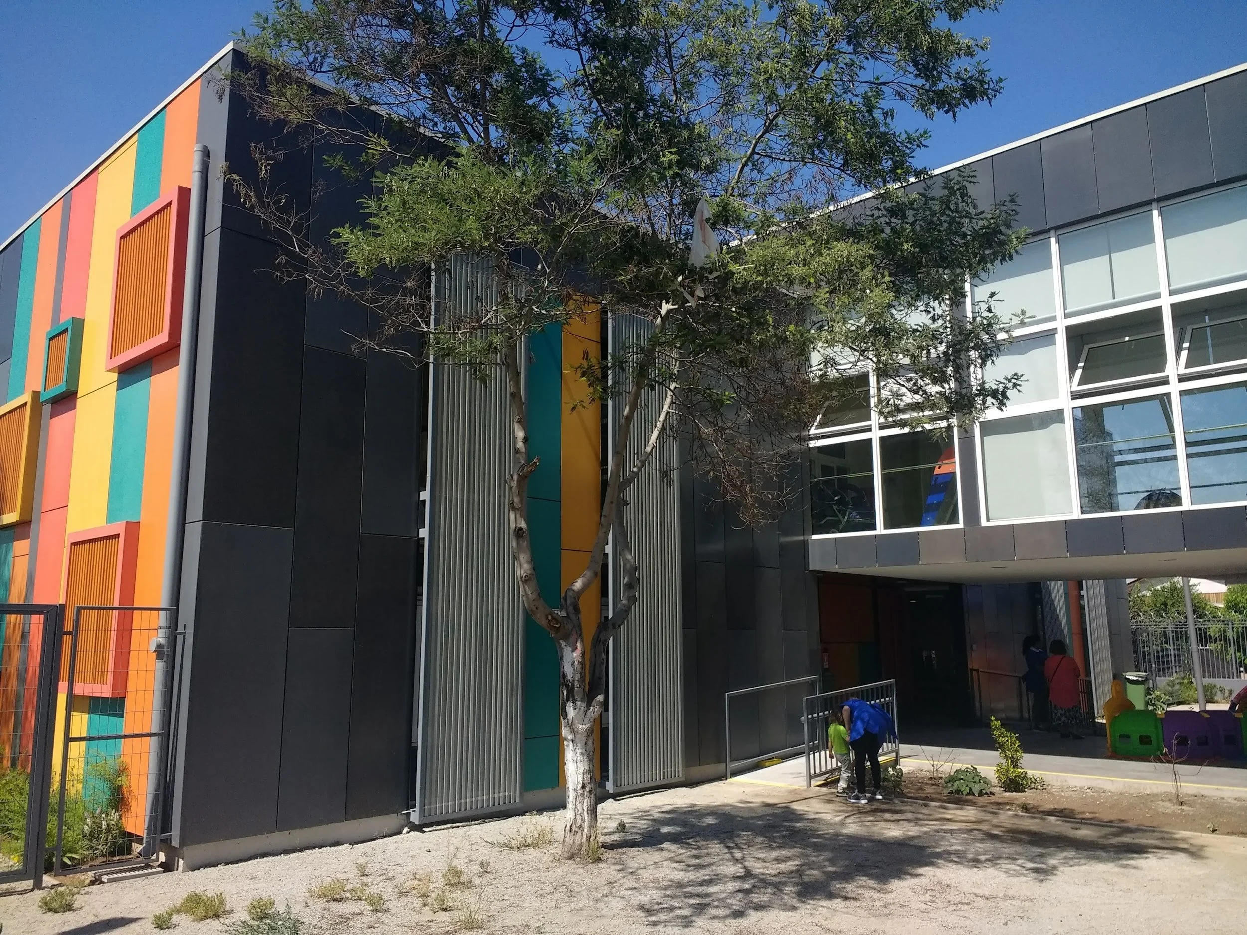 Edificio moderno con paredes negras y detalles de colores vibrantes en las ventanas, con árboles y niños jugando en el patio exterior.