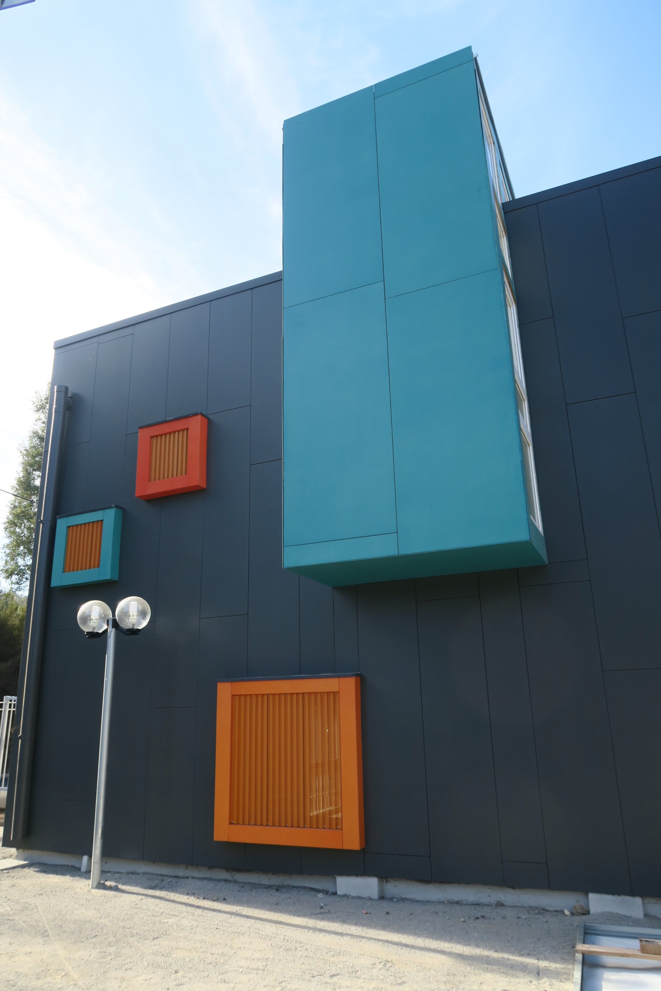 Edificio moderno con paredes negras, ventanas de madera naranja y elementos en azul y naranja. En el primer plano hay una farola y en el fondo se ve cielo despejado y algunos árboles.