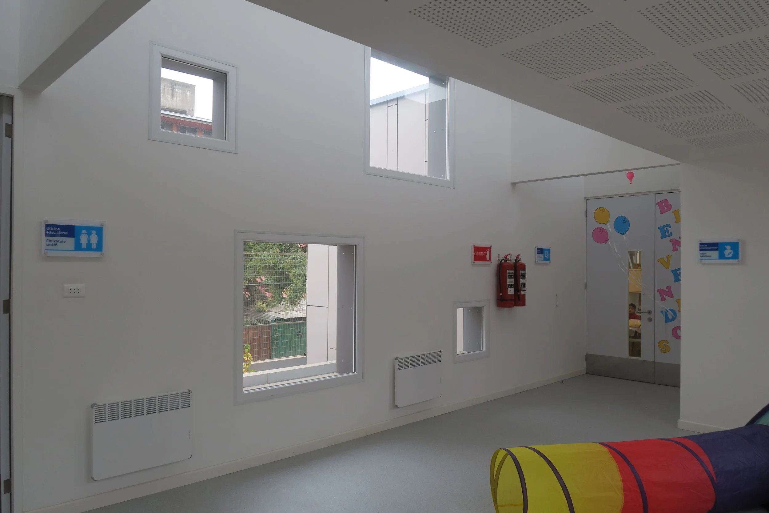 Interior de una escuela infantil con paredes blancas, ventanas altas y un área de juegos con puerta decorada con letras y globos de colores.