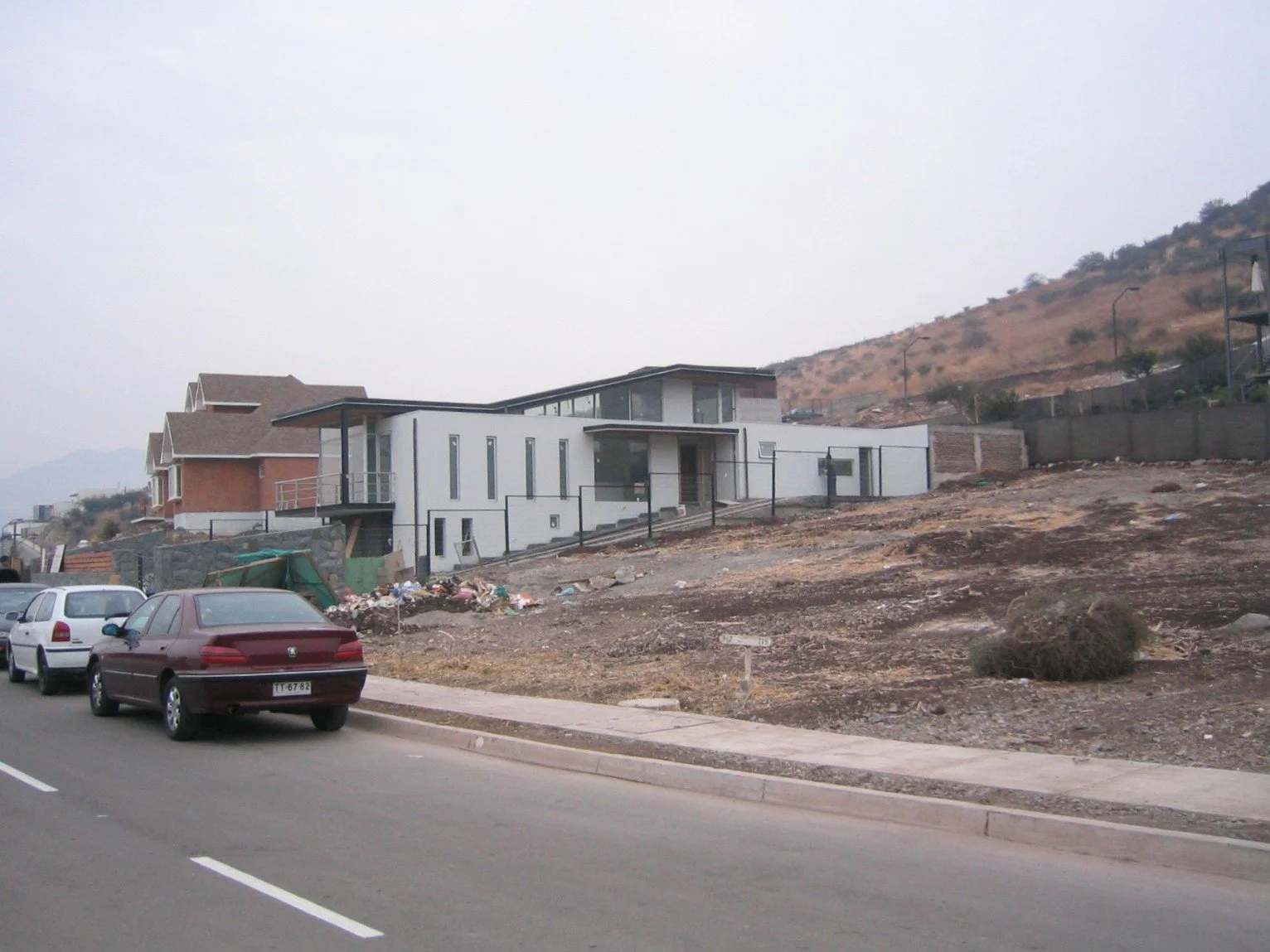 Casas modernas en una zona en construcción con vehículos estacionados en la calle, en un terreno árido y colinas al fondo.