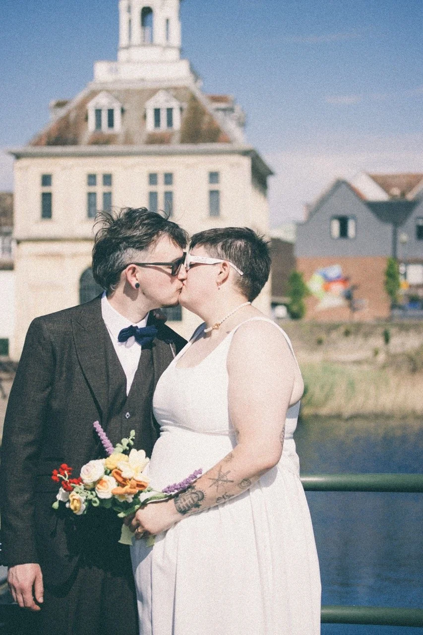 A same-sex couple kissing, holding a bouquet of flowers, outdoors near water with houses and a large historic building in the background.