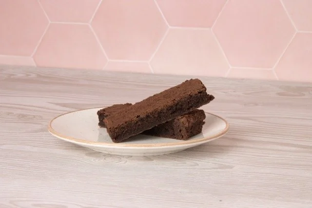 Two chocolate brownies on a white plate with a light wood surface and a pink hexagon tile wall in the background.