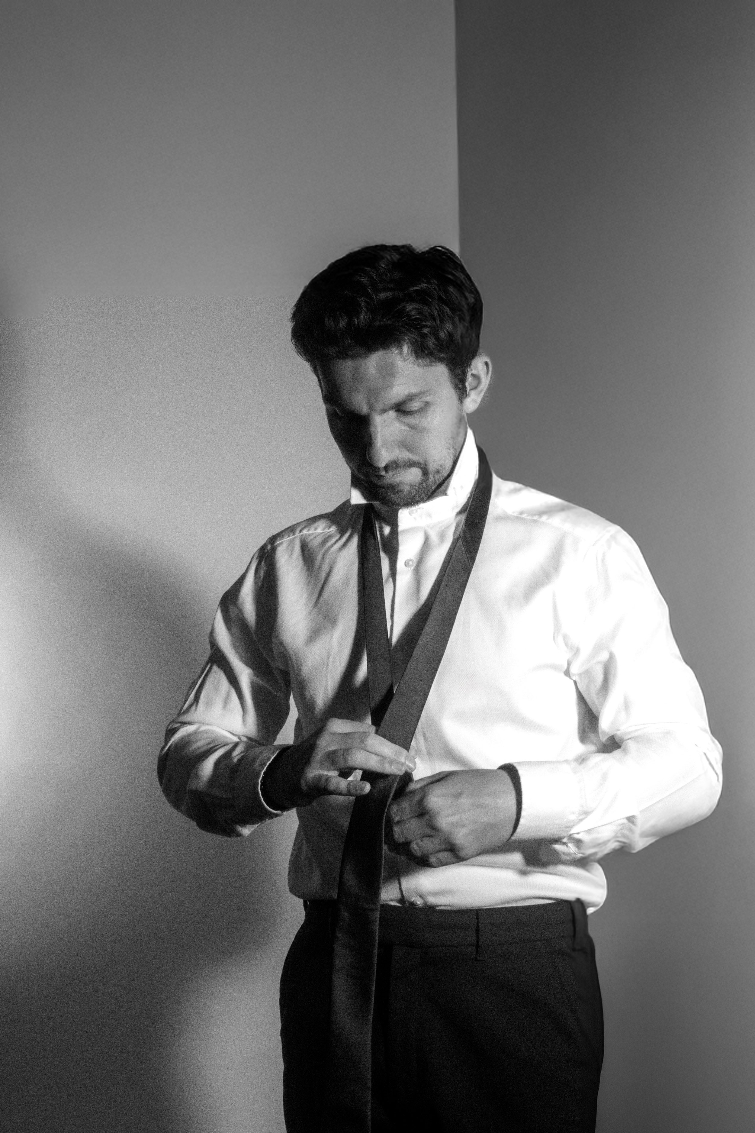 A man in dress shirt and pants adjusting his tie in front of a plain wall, with a serious expression and shadowed face.