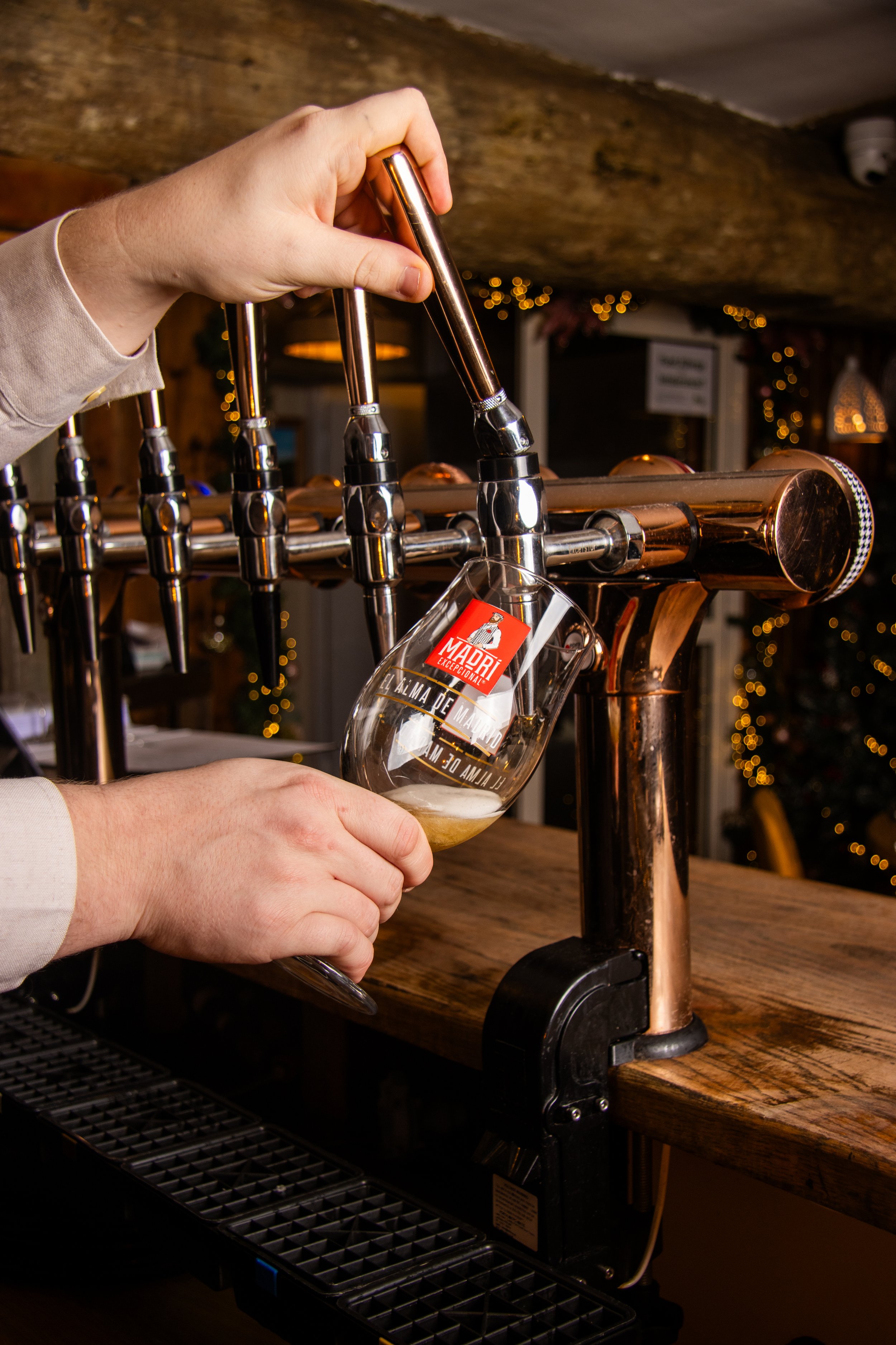 Person pouring beer from tap into glass at bar counter with warm lighting in background.