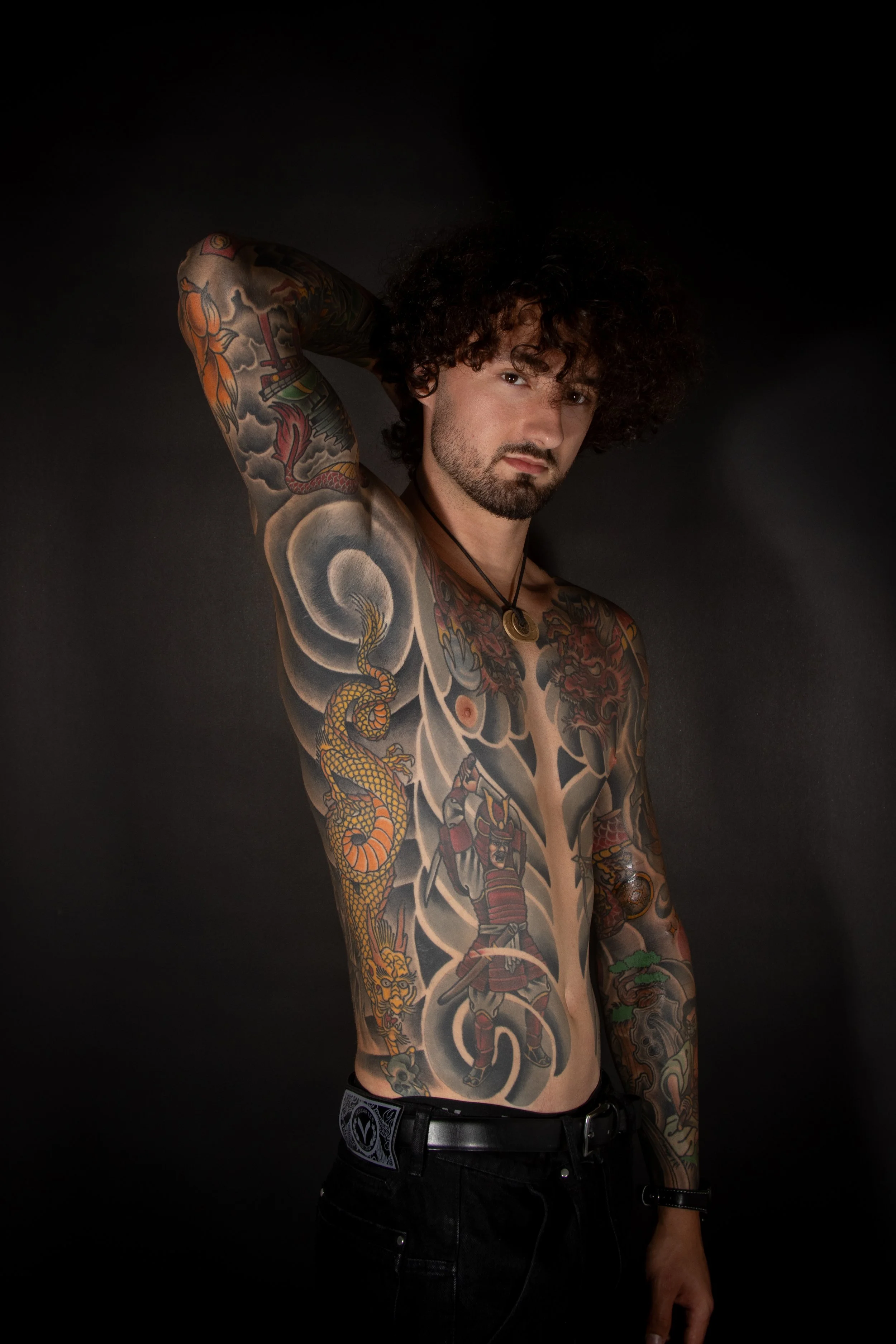 A studio photograph of a shirtless man with curly hair and tattoos covering his chest and arms, posing against a black background.