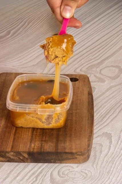 Person dipping a piece of bread into a container of caramel sauce on a wooden cutting board.