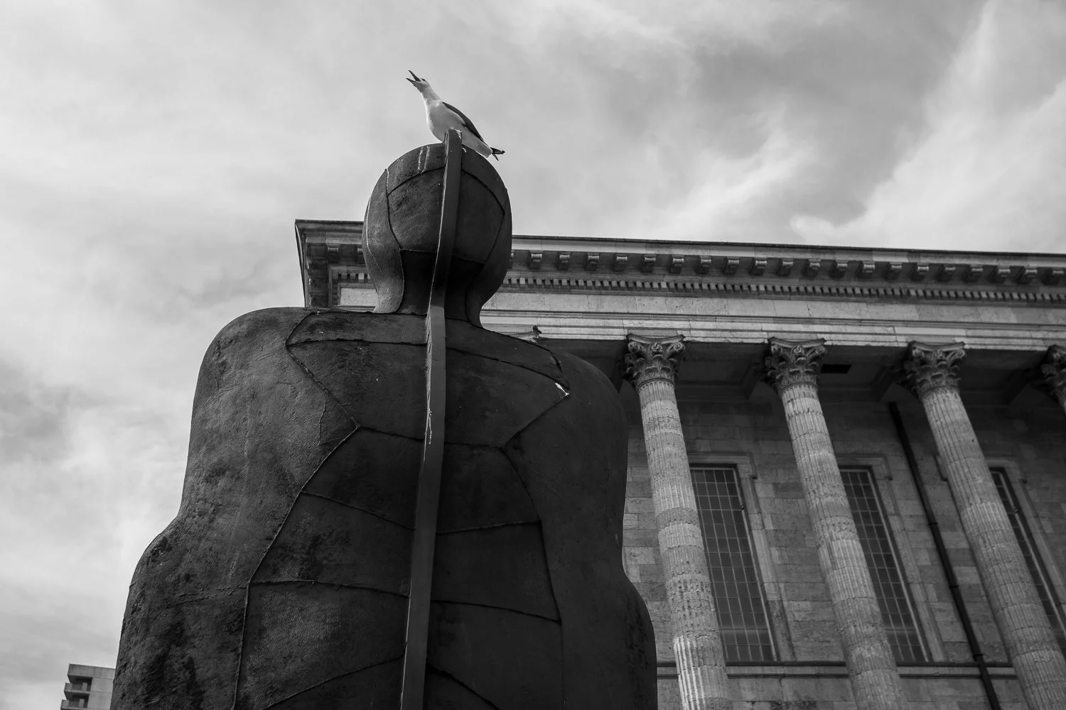 Black and white photograph of the Iron:Man sculpture in Birmingham with a small bird perched on its head, blending urban art with a touch of nature.