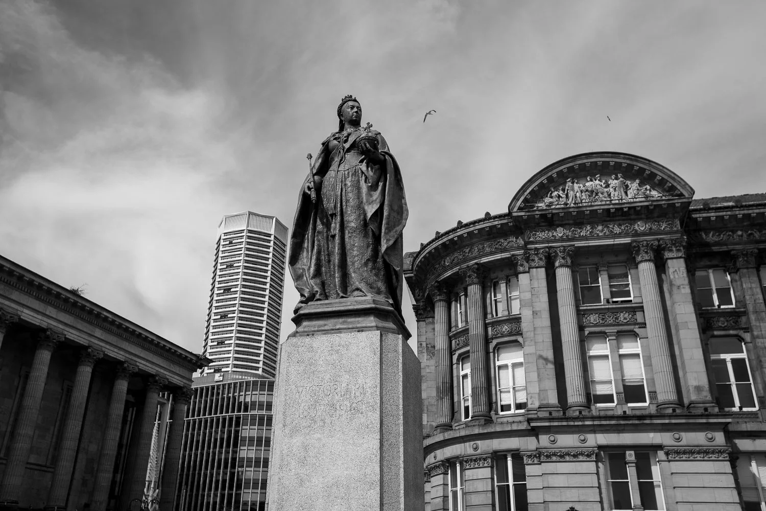 Black and white photograph of the Queen Victoria statue in Victoria Square, Birmingham, showing historic sculpture with classical buildings in the background.