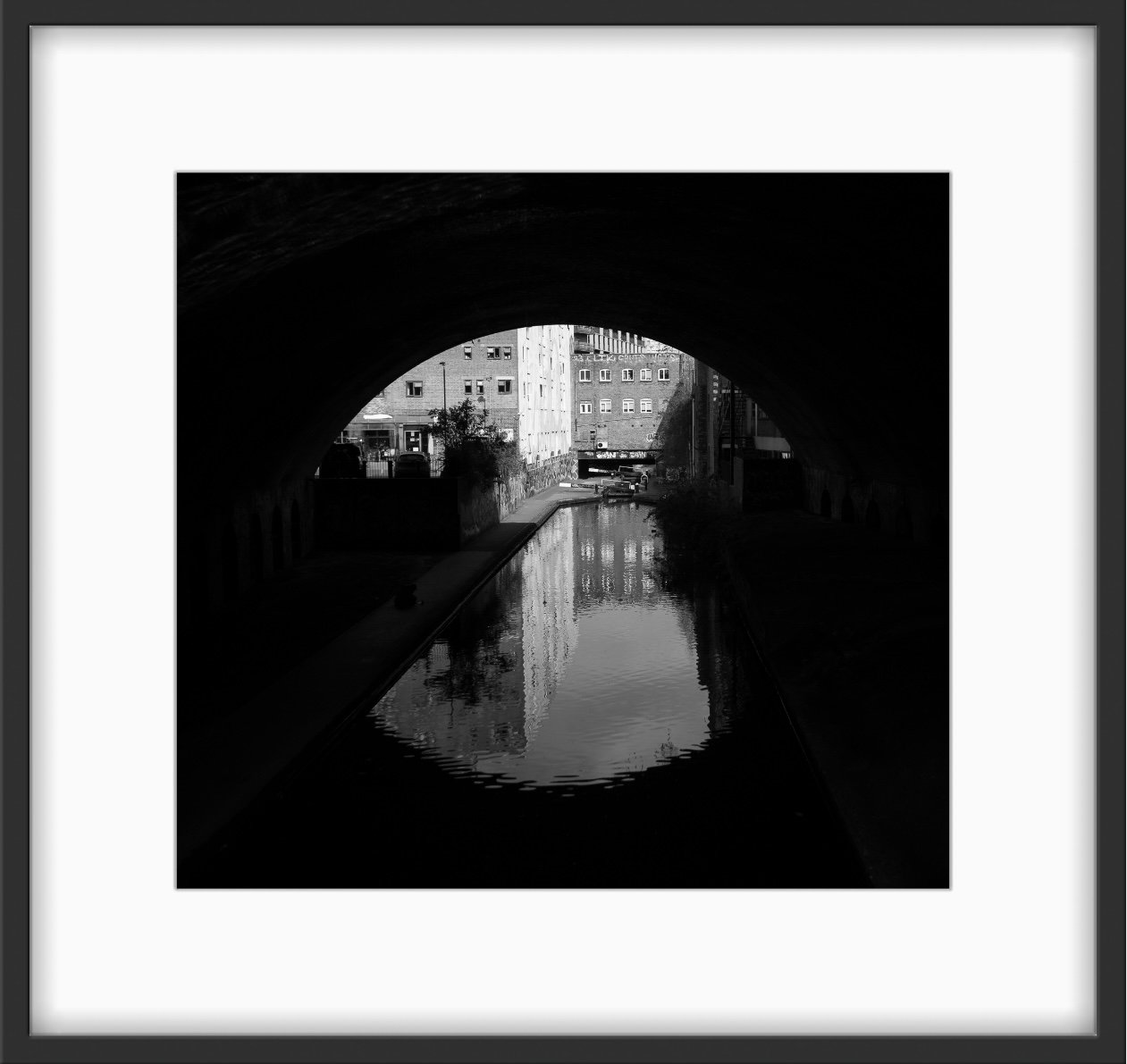 Square black and white photo of Birmingham canal seen through a tunnel, showing a Victorian industrial building with faint graffiti and soft reflections on the water.