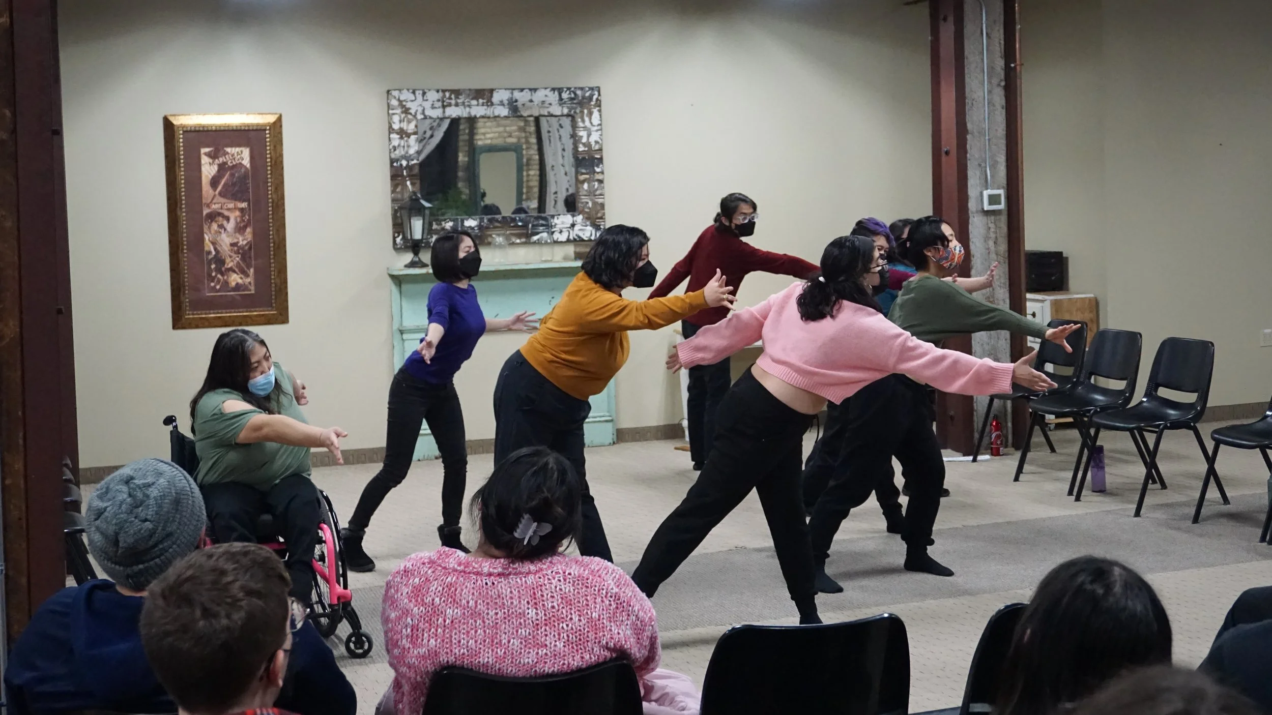 A group of BIPOC performers in black pants, colored shirts, and masks reach across in the same direction