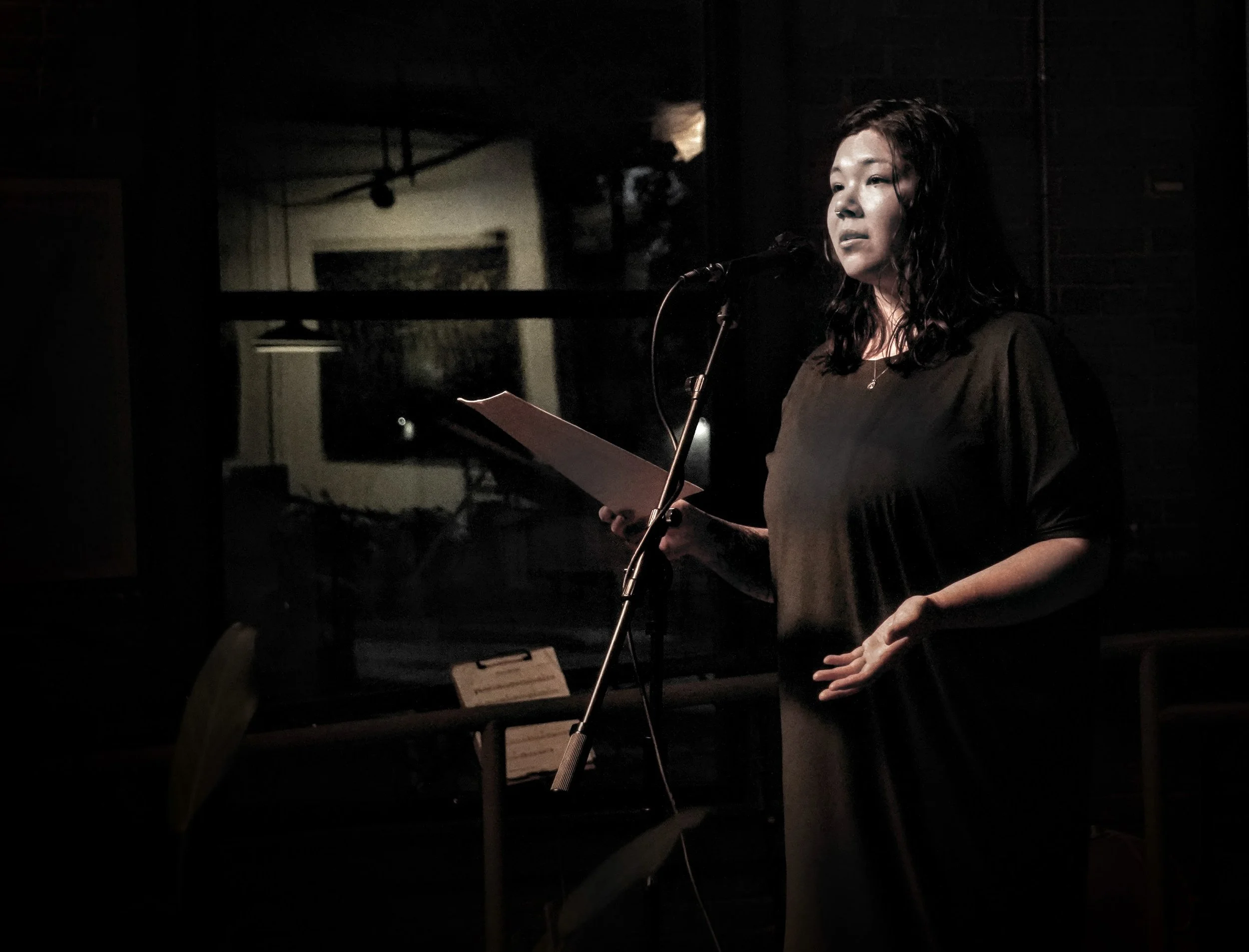 Pauline in a dress with her open palm in front of her, reading from papers into a microphone in dim lighting