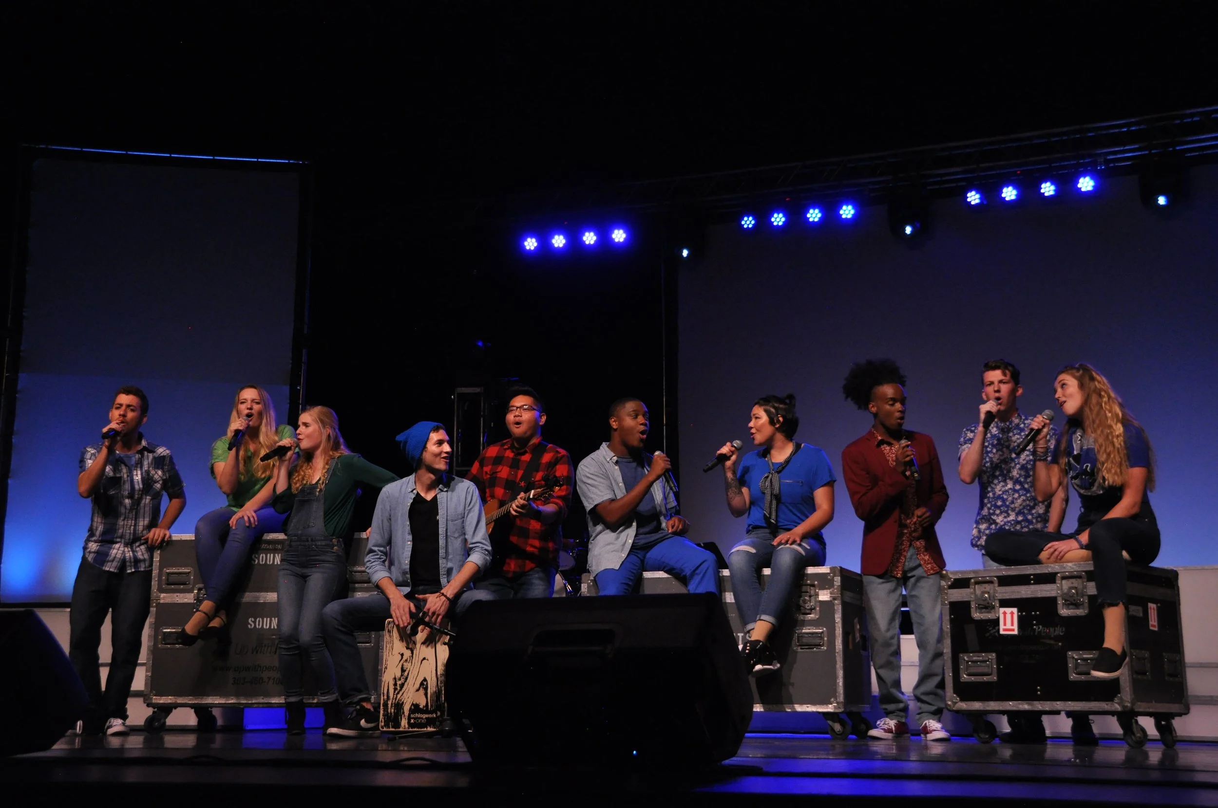 Pauline and nine other performers sit onstage on rolling equipment boxes. Pauline and seven others sing into microphones with one drummer and one guitarist.