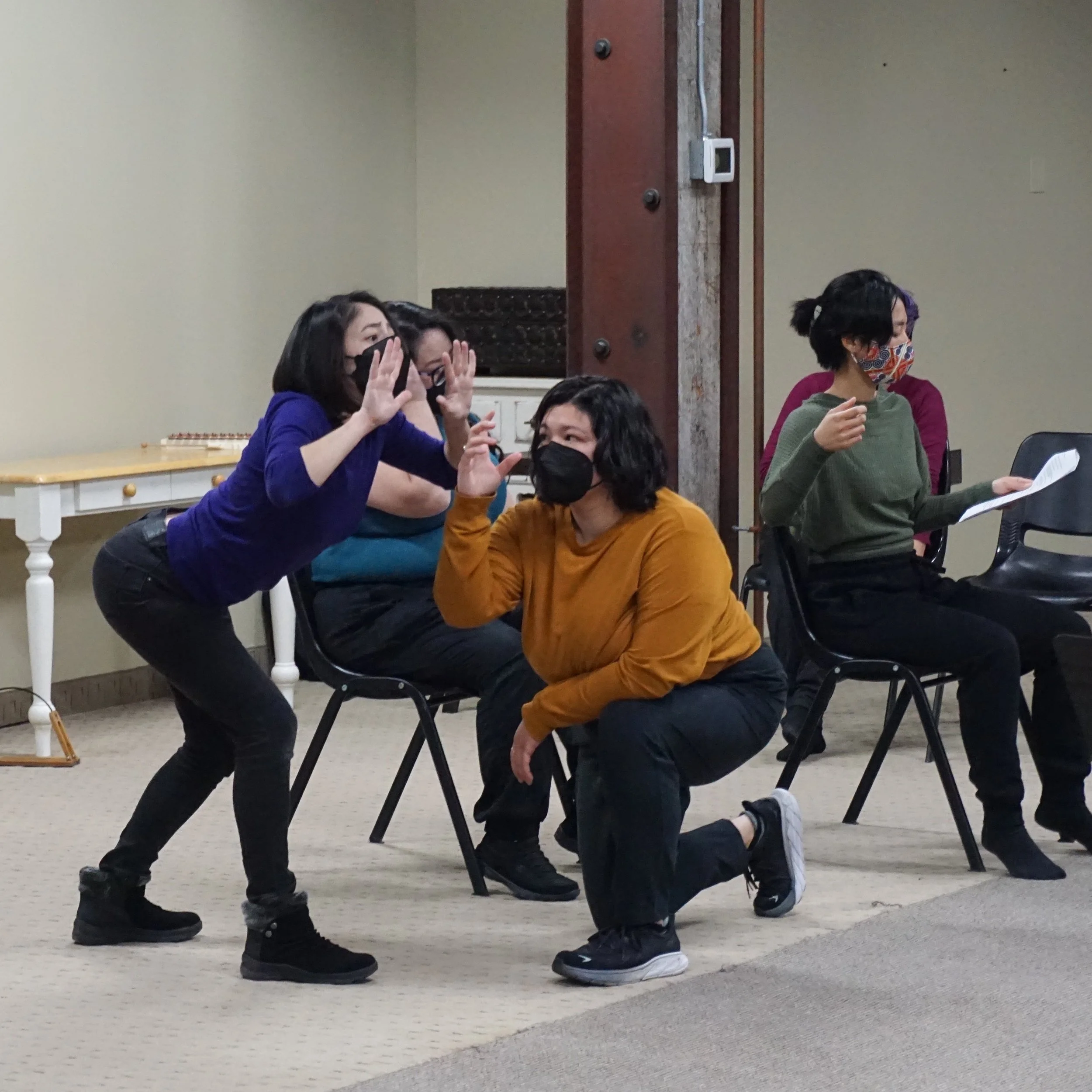 Pauline in black pants, a pumpkin-colored shirt, and a black KN95 respirator crouched on one knee looking at a fellow performer