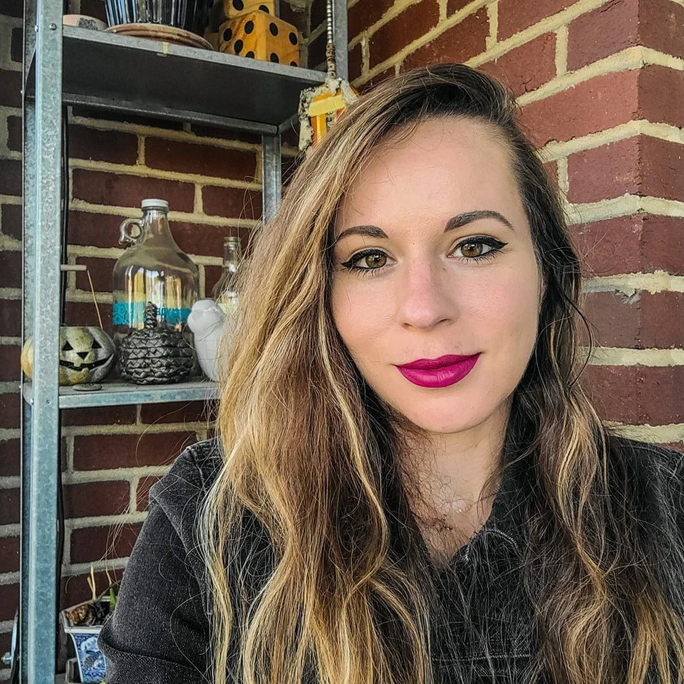 A woman with long wavy hair and bright lipstick taking a selfie outdoors against a background of a brick wall and shelves with decorative items.