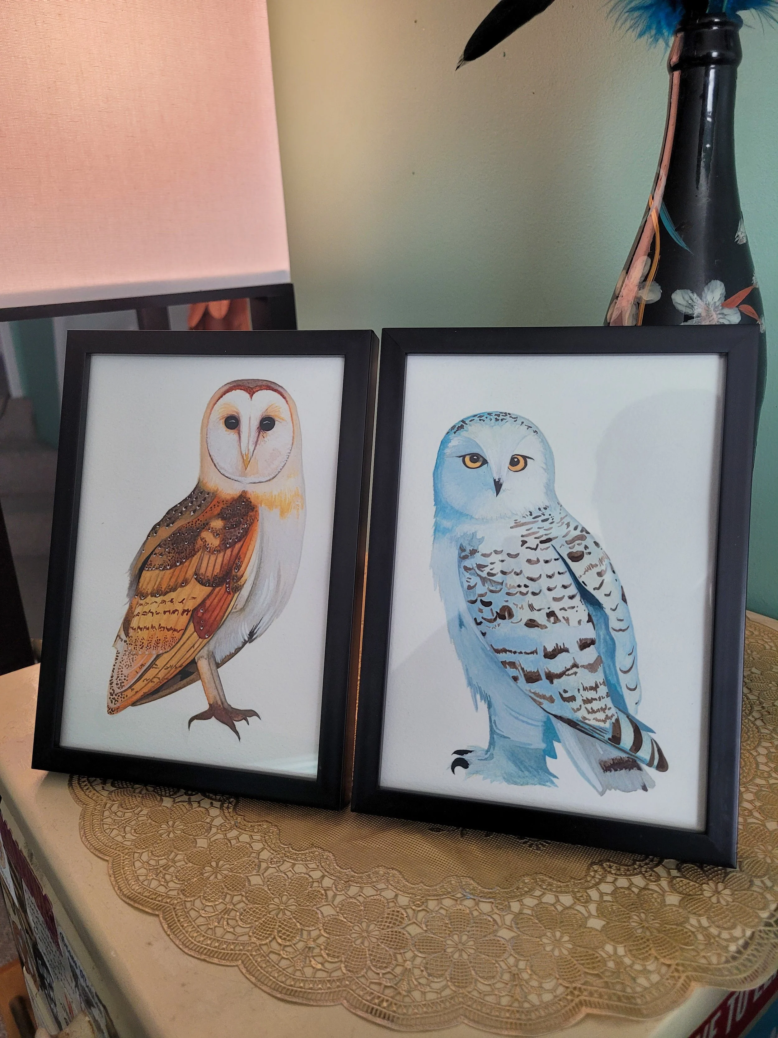 Framed watercolor paintings of an owl and a snowy owl on a table with a lace tablecloth, beside a lamp and a floral vase.