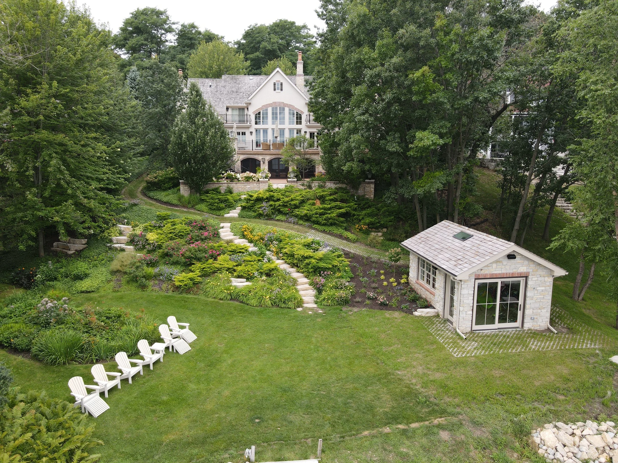 Lush backyard with green lawn, a landscaped garden with colorful flowers, a winding stone pathway, several white Adirondack chairs, and a small stone shed. In the background, a large multi-story house with large windows and balconies is surrounded by tall trees.