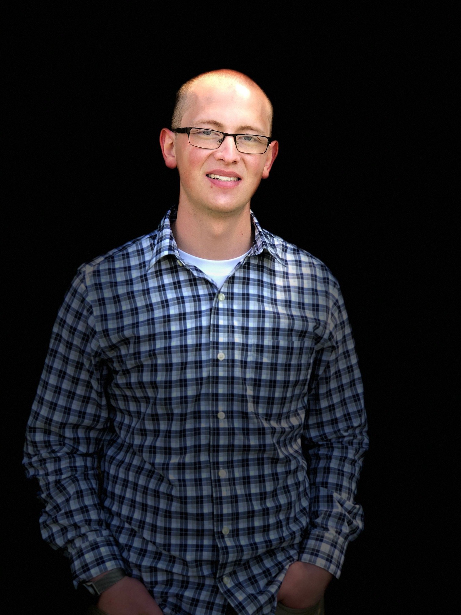 A young man with glasses, short light hair, and a slight smile, wearing a blue and white checkered shirt, standing against a black background.