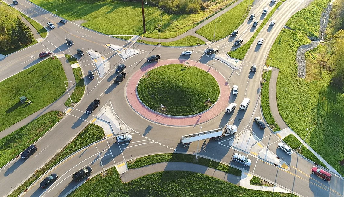 Aerial view of a roundabout with multiple roads connecting, cars circulating, and green grassy areas in the center and along the sides.