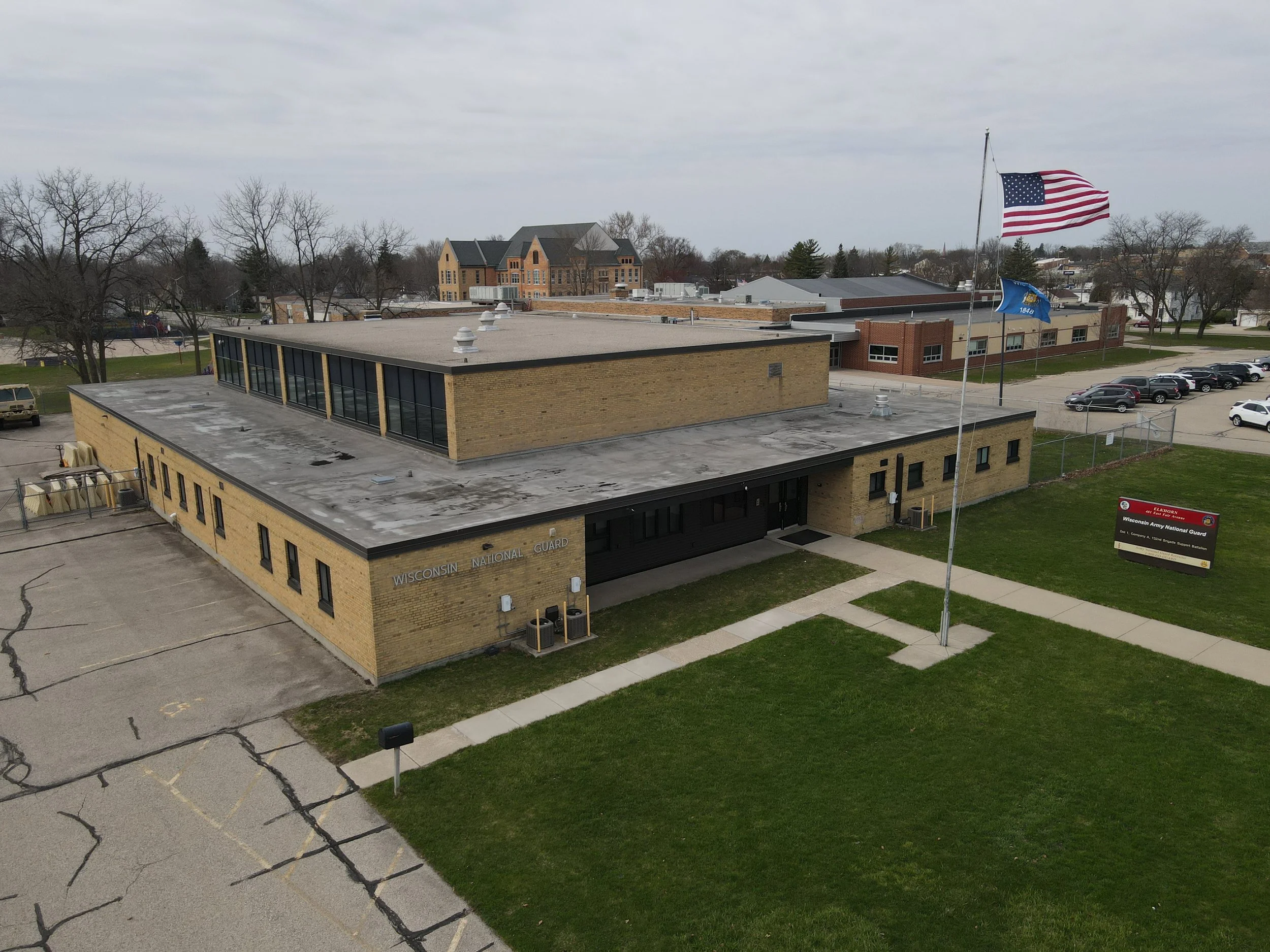 Aerial view of a brick building with a sign that reads 'Wisconsin National Guard' and a flagpole with the American flag and Wisconsin state flag. The building is surrounded by a parking lot, grass, and a sidewalk.