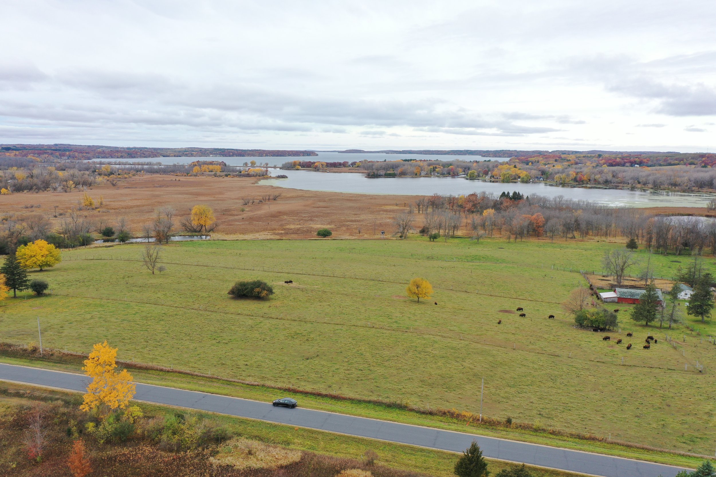 A rural landscape with a grassy field, trees with fall foliage, a winding road with a black car, a farm with red barn and white house, and a river in the background under cloudy sky.
