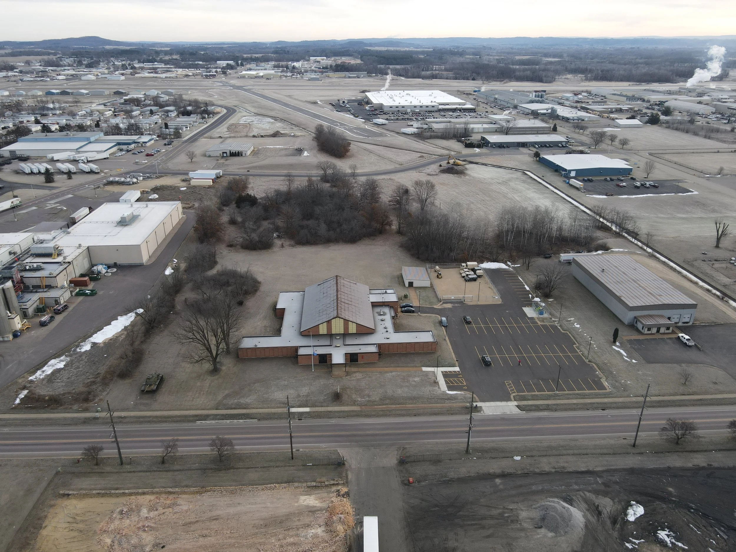 Aerial view of a semi-urban area with a church and parking lot in foreground, industrial buildings and warehouses in the background, and a parking lot with a few cars.