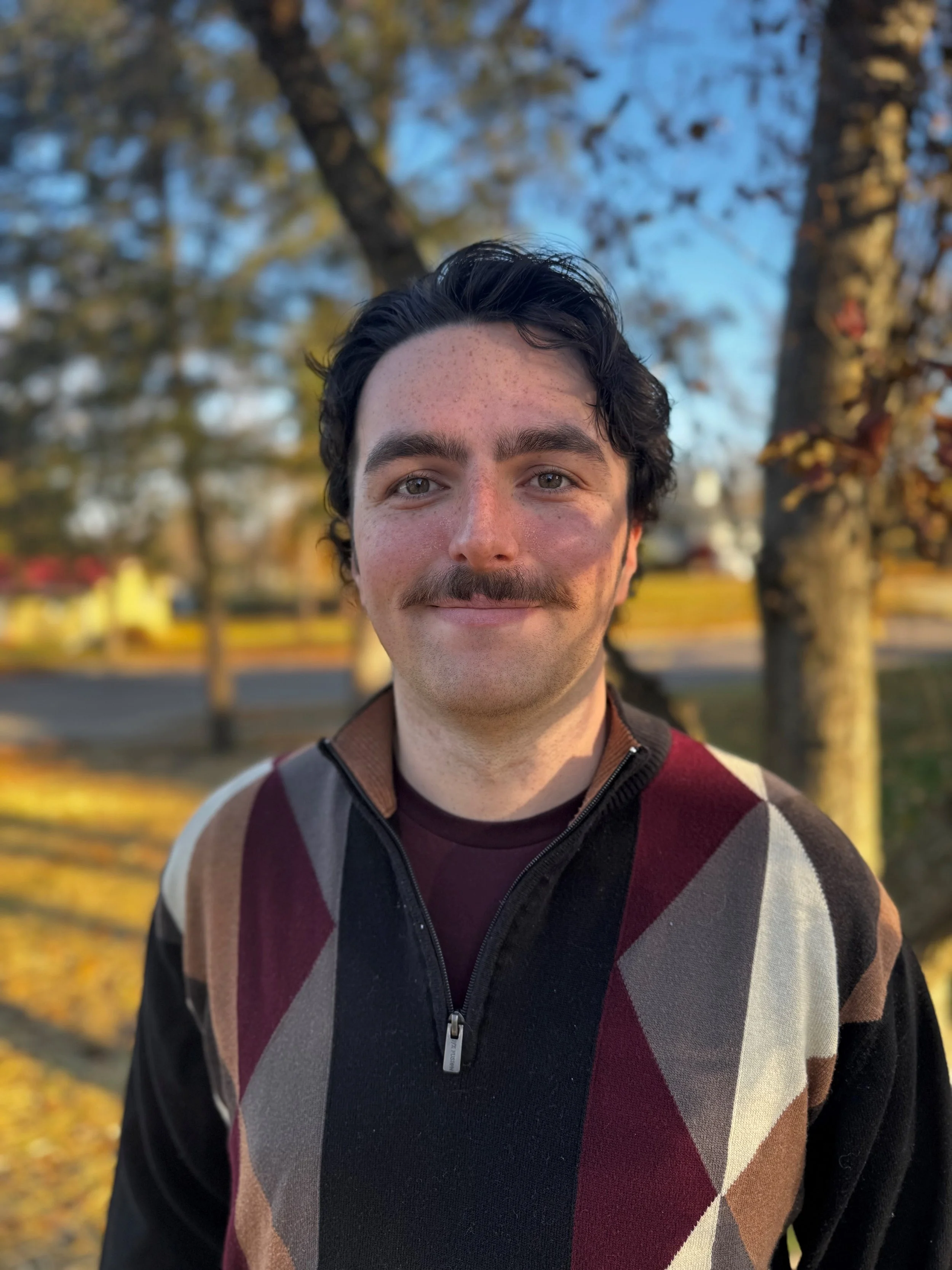 A young man with dark, curly hair and a mustache, smiling outdoors in front of autumn trees, wearing a multicolored patterned sweater.