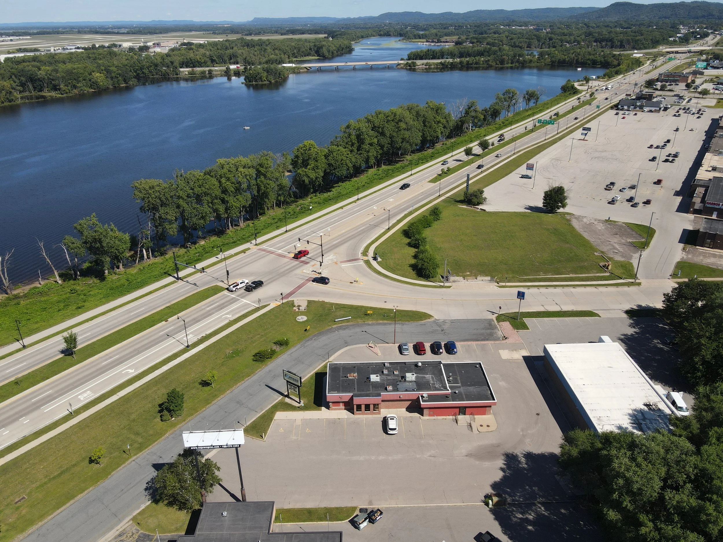 Aerial view of a river, a busy road with cars, and commercial buildings with parking lots, surrounded by green trees and grass.