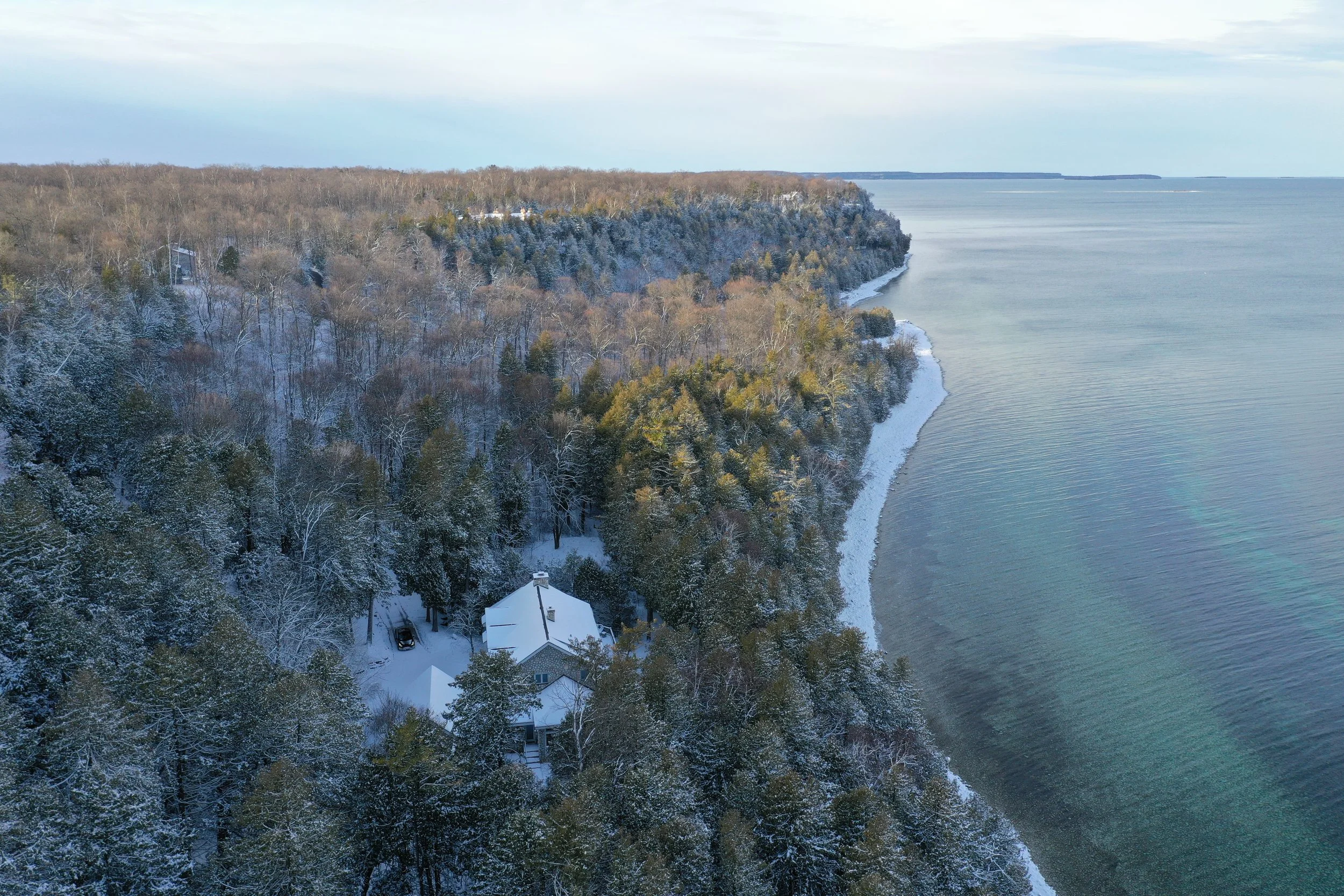 A snow-covered coastal landscape with a house surrounded by trees near the shoreline and a shoreline extending into the distance under a cloudy sky.