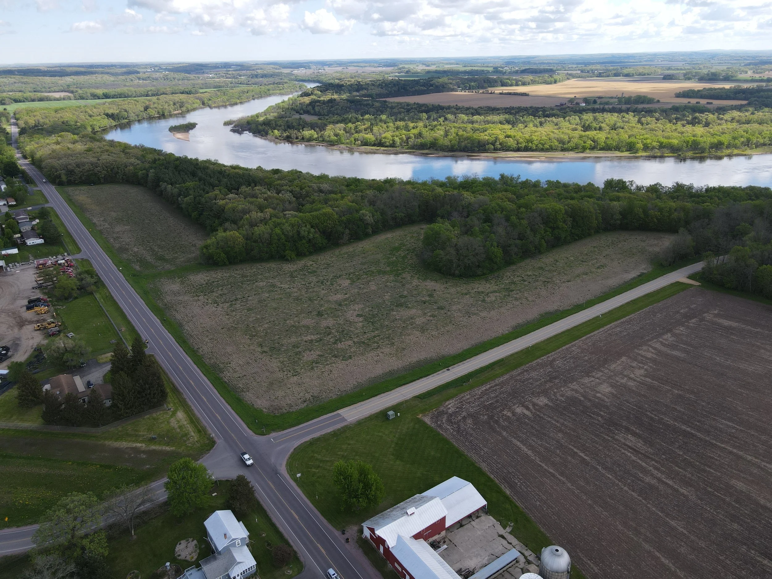 Aerial view of a rural landscape with a winding river, surrounded by lush green trees, farmland, a few houses, and roads.