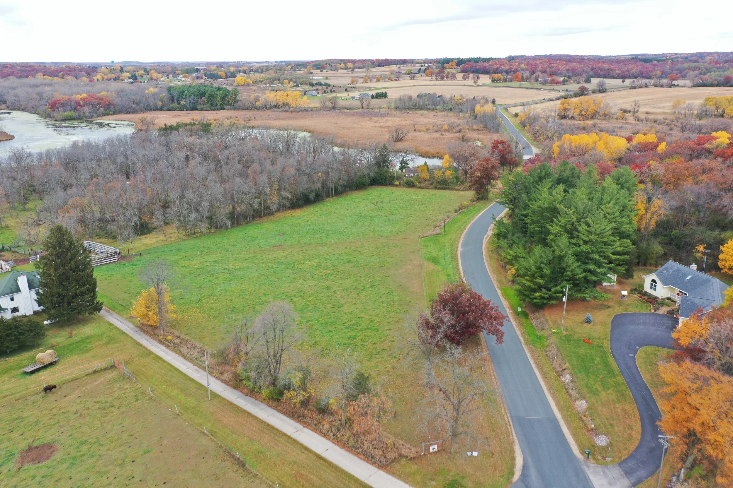 Aerial view of a rural landscape during fall, showing a curved road, green and leafless trees, a grassy field, houses, and a distant river with colorful trees.