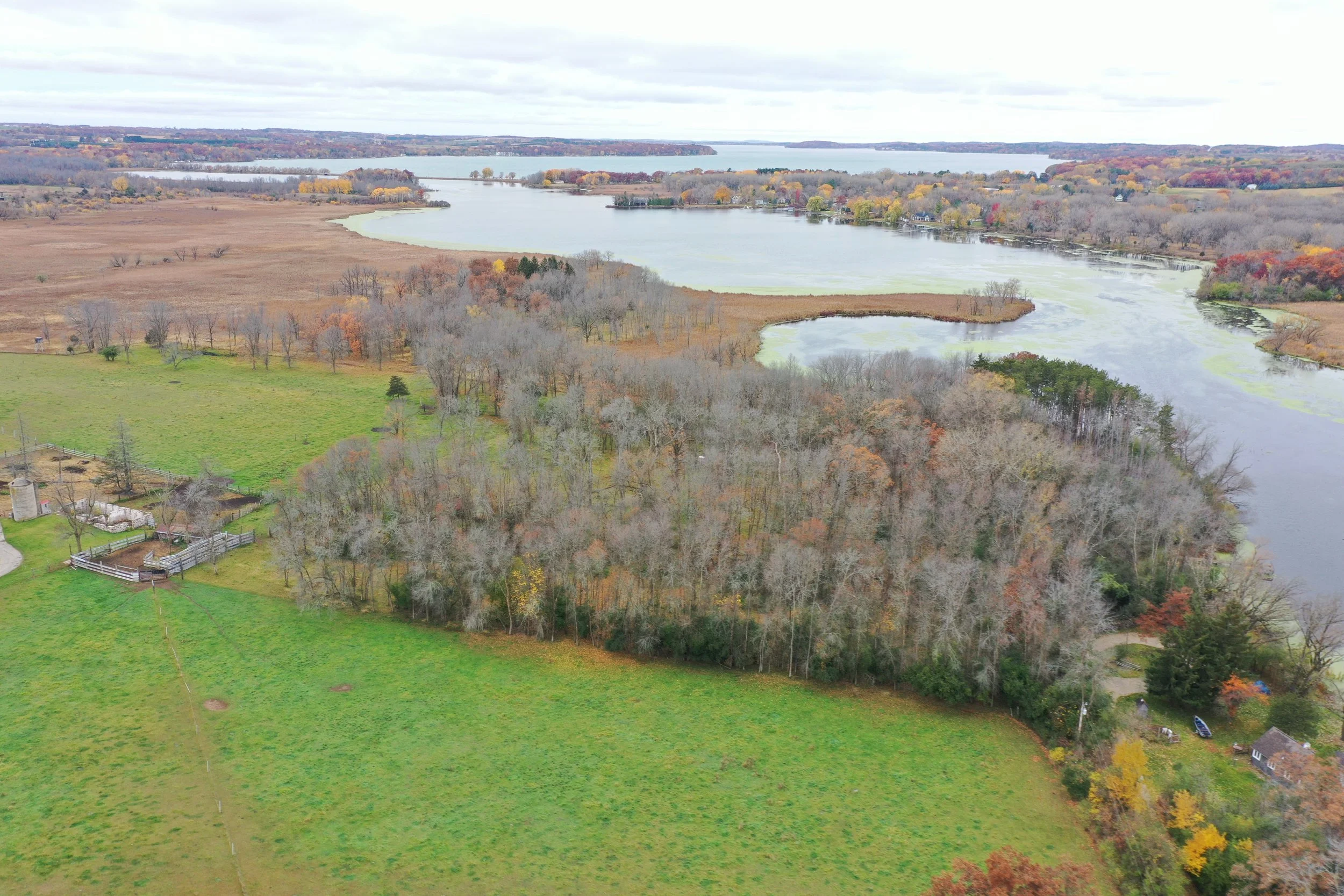 Aerial view of a landscape featuring a river, wooded area, grassy fields, and a few houses in autumn with colorful trees.