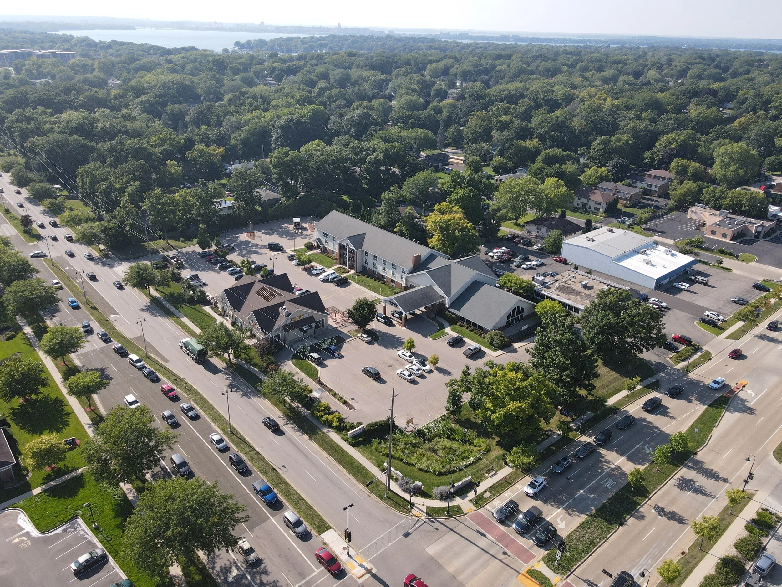 An aerial view of a commercial and residential area with parking lots, buildings, and tree-lined streets on a sunny day.