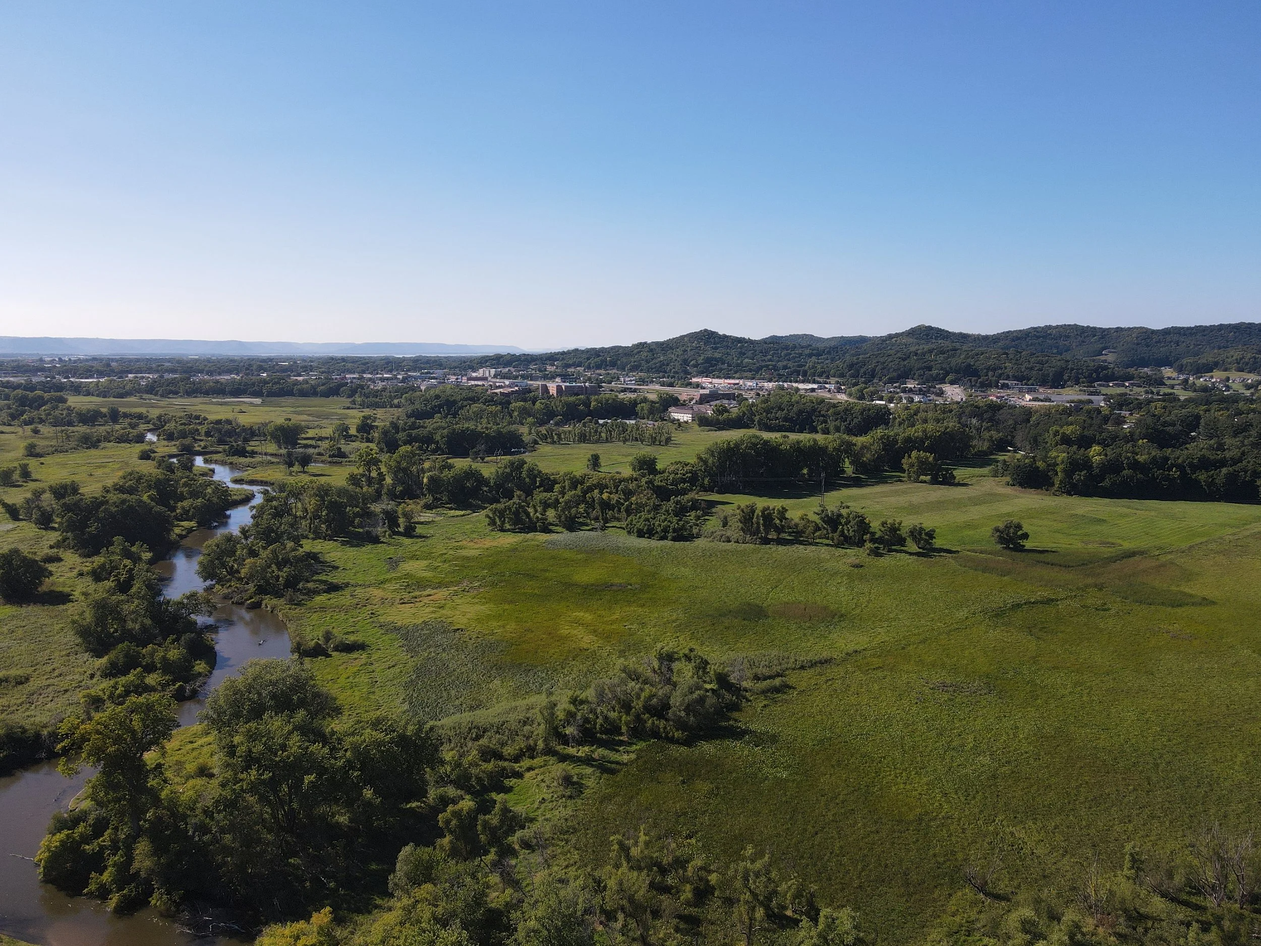 Aerial view of a lush green landscape with a winding river and trees, with a town and hills in the background under a clear blue sky.