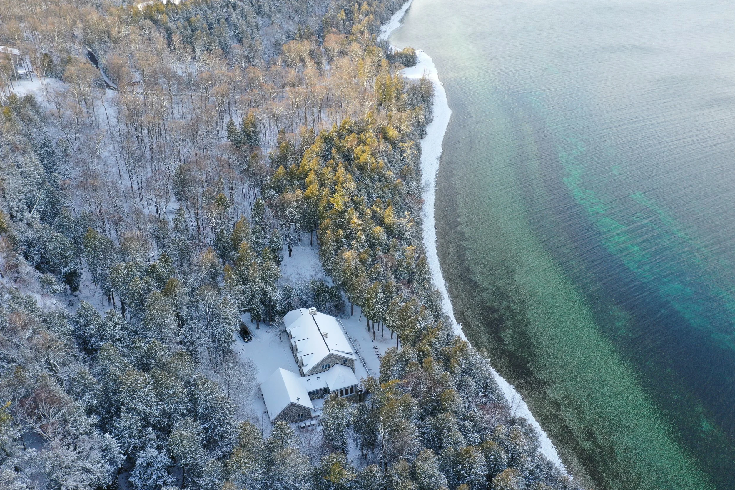 An aerial view of a snowy landscape with a house surrounded by trees and a shoreline, next to a body of water with visible underwater features.