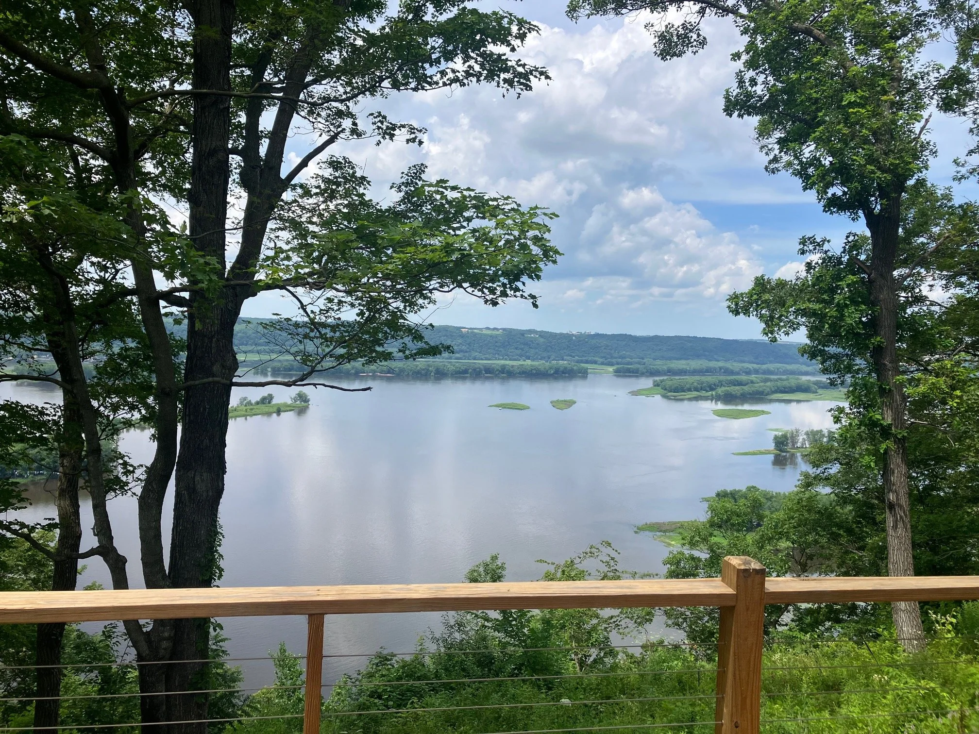 Scenic view of a river with small islands, framed by trees and a wooden railing in the foreground, under a partly cloudy sky.