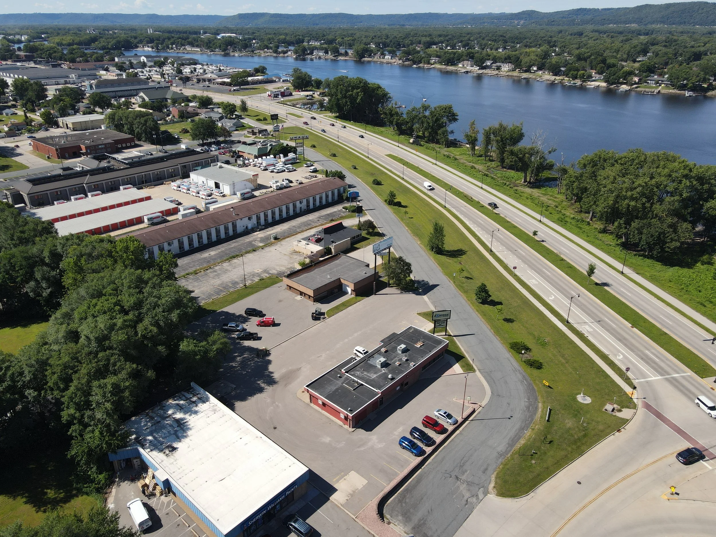 Aerial view of a commercial area near a river, showing parking lots, buildings, roads, trees, and a large water body in the background with hills.