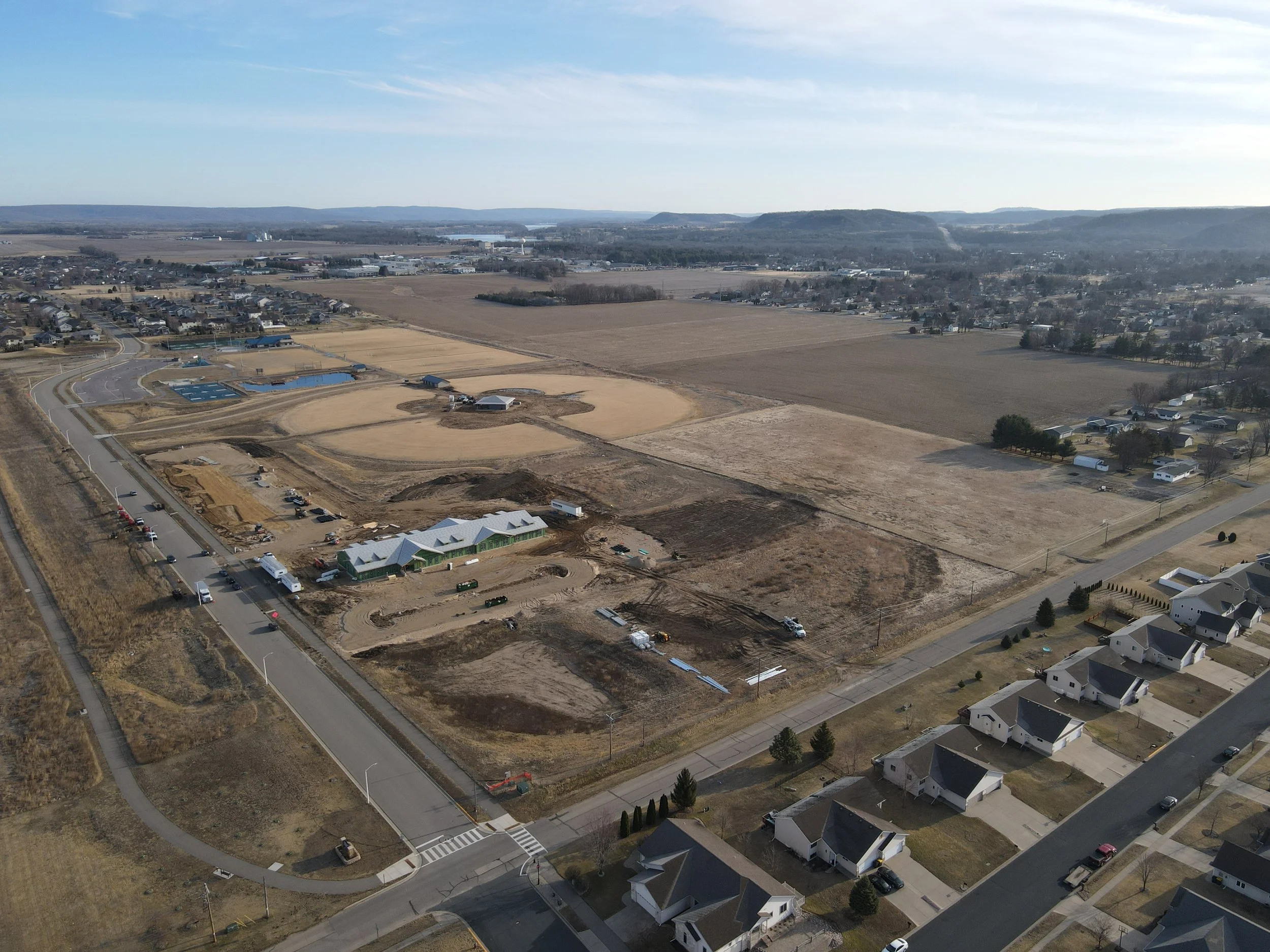 Aerial view of a suburban area under construction, with several buildings, roads, and residential houses, and open fields.