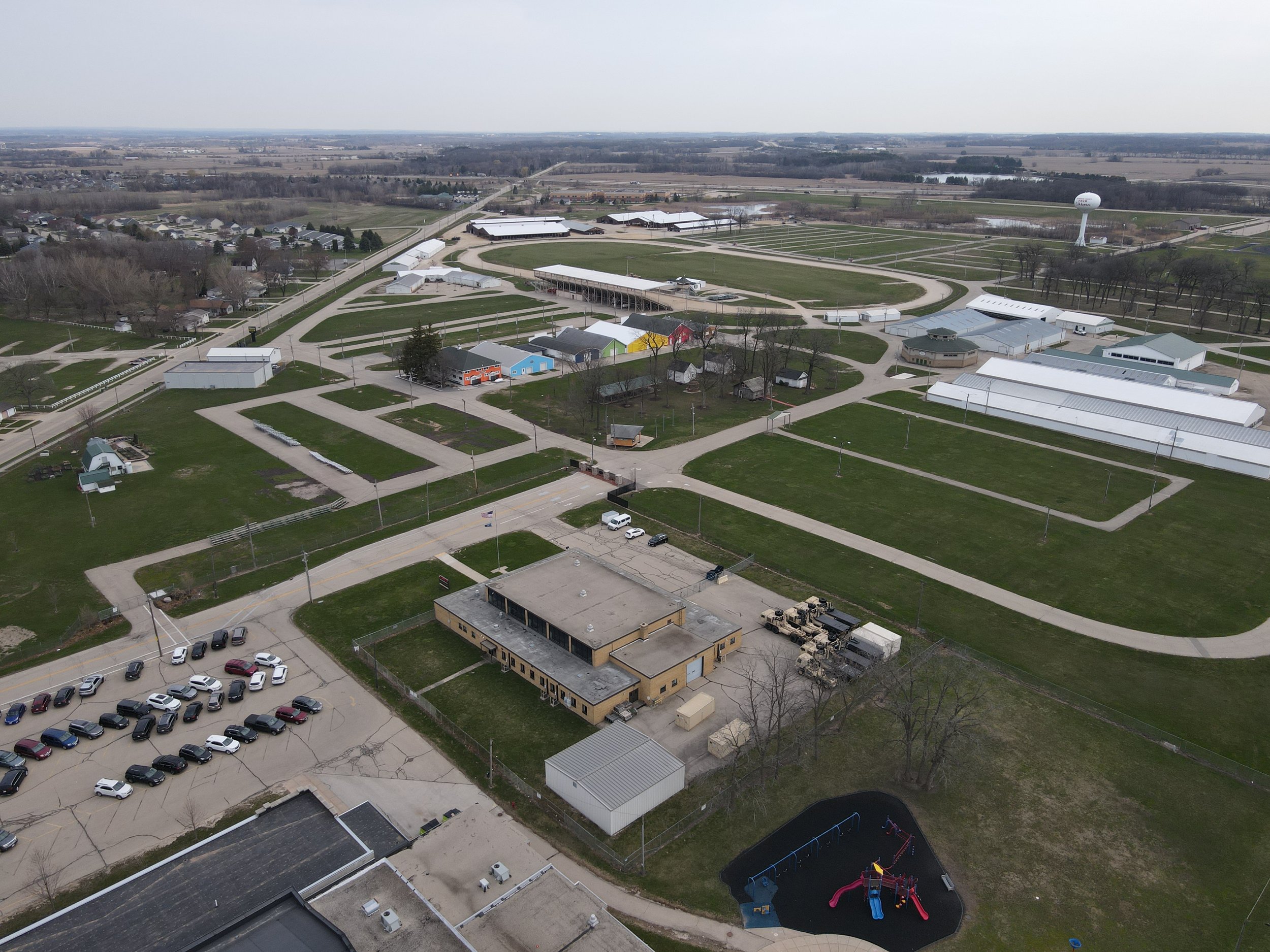 An aerial view of a large park with green fields, pathways, and various buildings. There is a playground with slides and swings, a parking lot with cars, and a water tower in the distance.