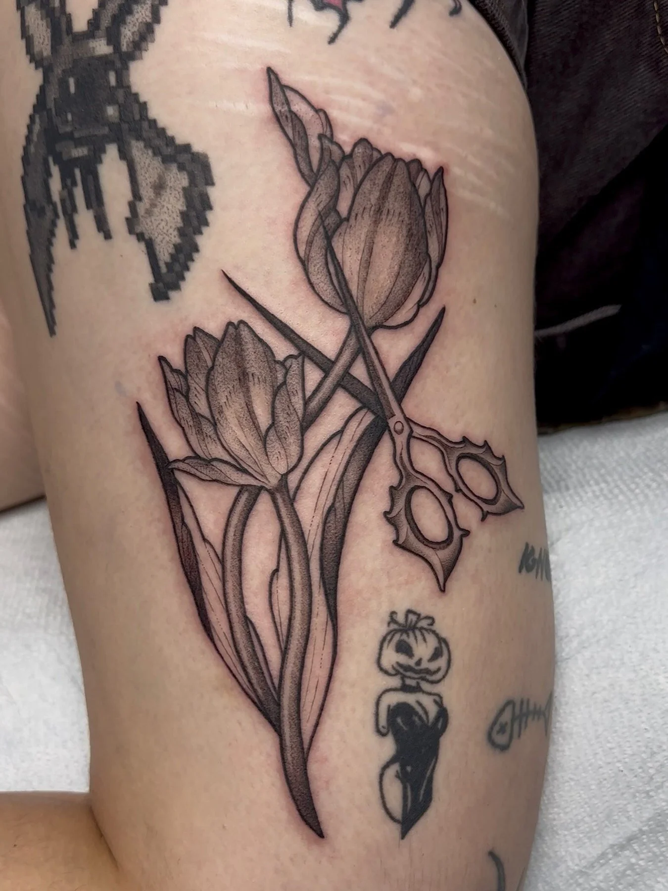A tattoo of tulips in outline on a person's upper arm, with a pair of scissors intertwined with the flowers and smaller tattoos below.